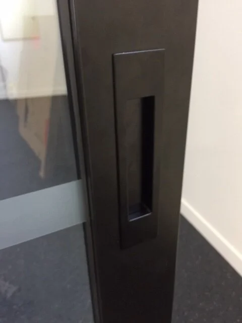 Close-up of a black metal door with a vertical rectangular handle and a narrow window.