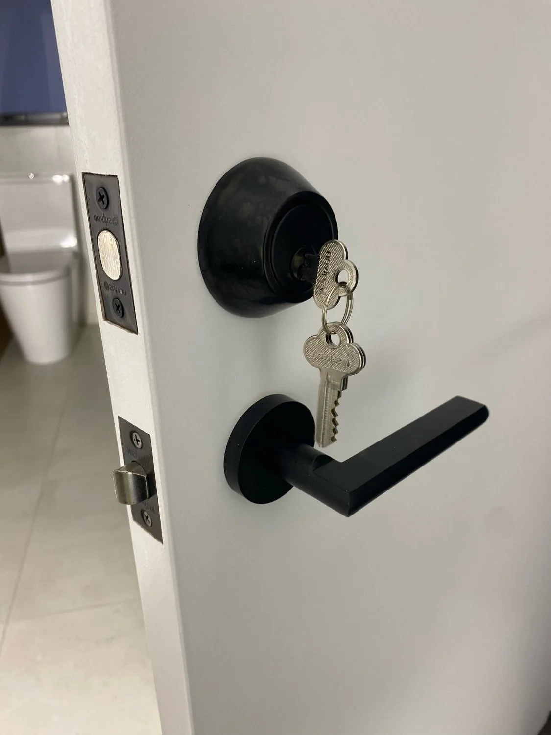 A close-up of a white door with a black handle and a key inserted into the door lock. The key is attached to a keyring with two small keys.