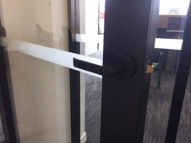 Clear plastic strip barrier on a glass door with a black handle and a small padlock, in an office setting.