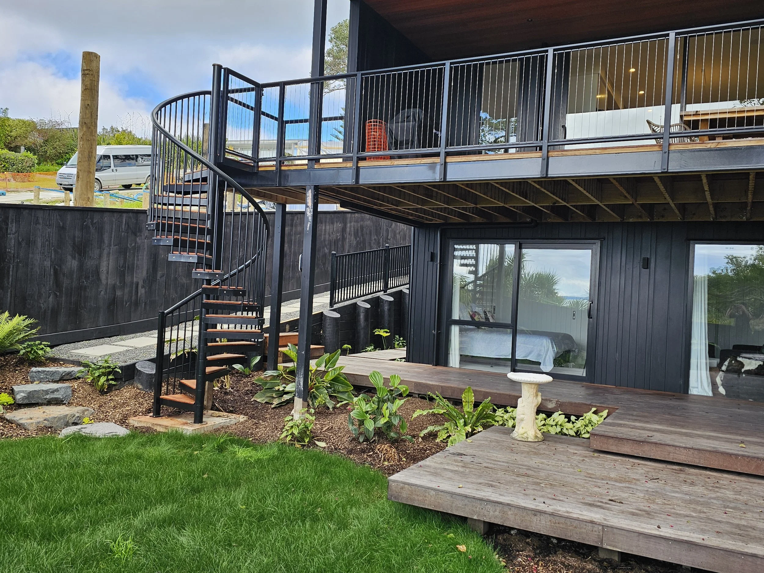 Modern two-story house with a black exterior, wooden deck, glass sliding doors, and a spiral staircase leading to a balcony with outdoor furniture, surrounded by a green lawn and landscaped garden.