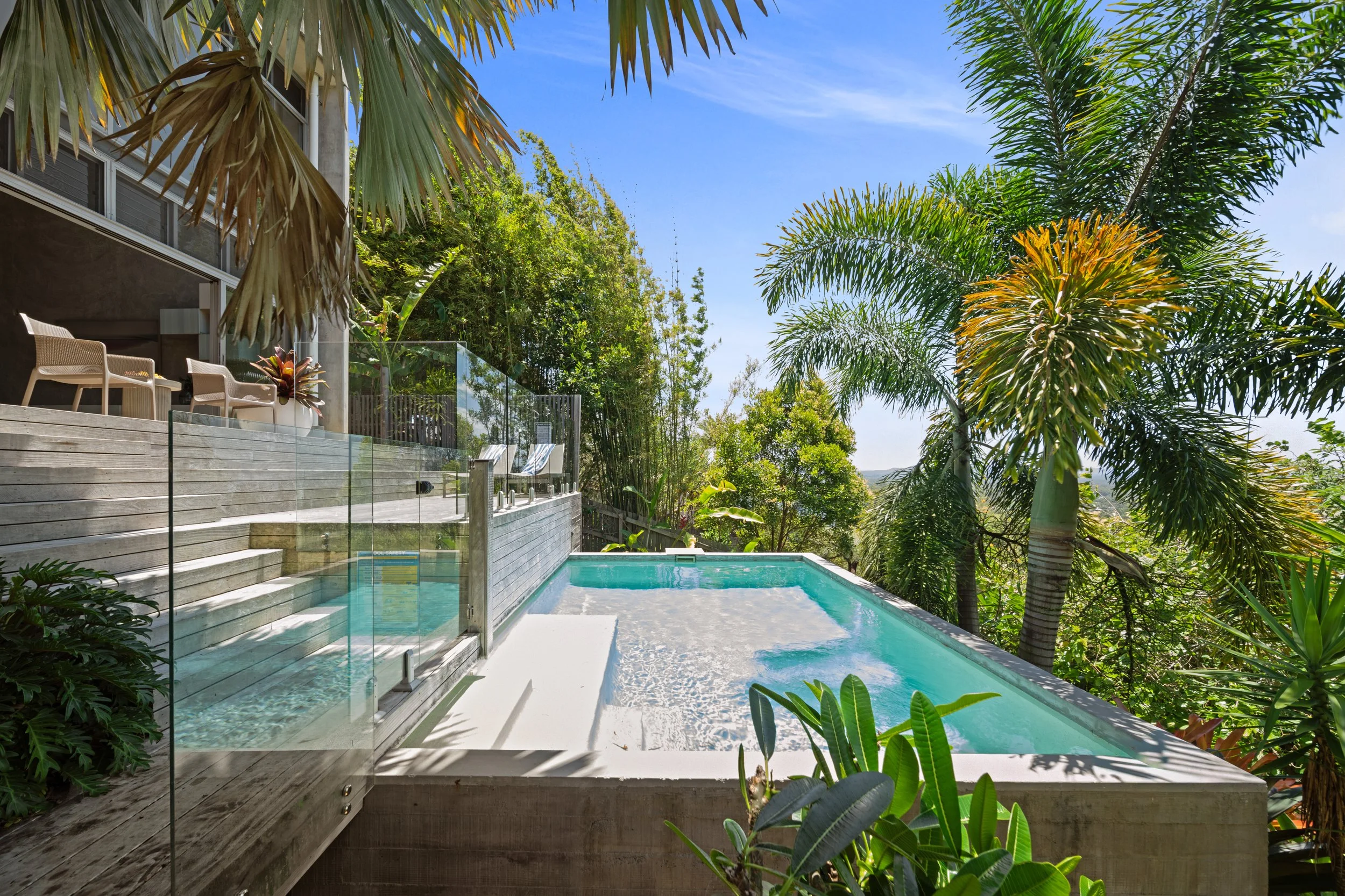 A backyard with a small pool, wooden deck, two white lounge chairs, a side table, and a glass safety fence. Lush green trees and hills are visible in the background under a partly cloudy sky.