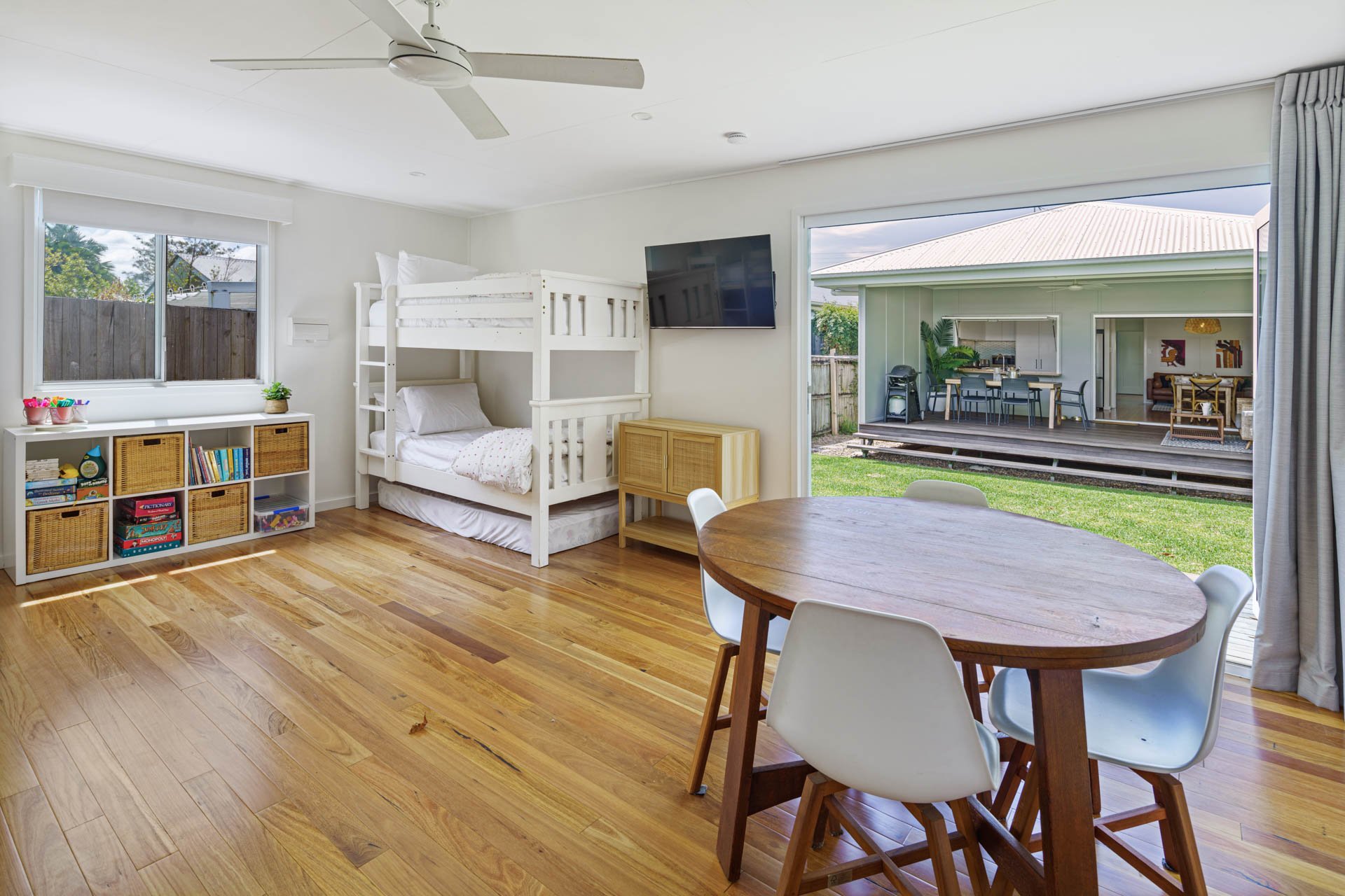 A woman standing on a porch looking into a dining room with a wooden table and chairs, with a dog sitting nearby. The porch has plants and a wooden floor, with the dining area visible through an open sliding door.