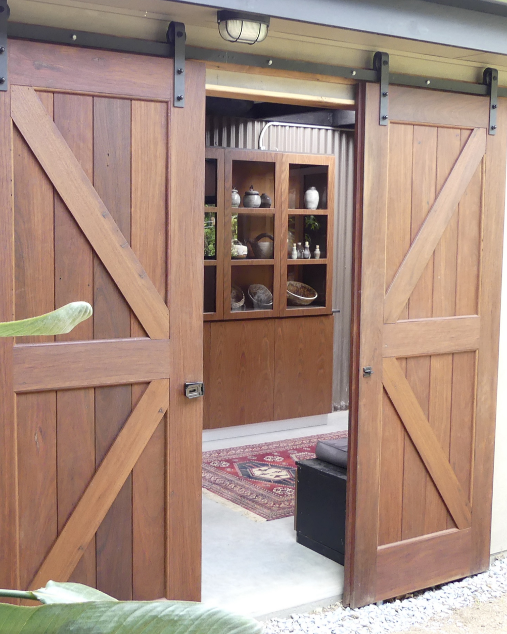 View of a room through wooden barn door style sliding doors, showing a wooden cabinet with glass doors, displaying various pottery and decorative items, and a patterned rug on the floor.