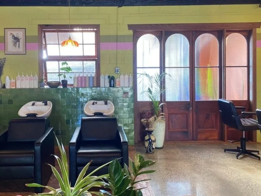 Interior of a vintage salon with two sinks, black chairs, green tiled walls, wooden door with glass panes, potted plants, and a window with a stained-glass lamp hanging above.