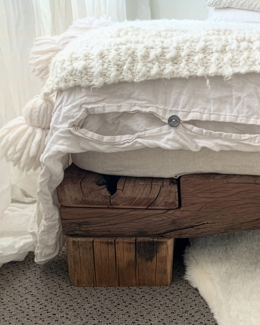 Close-up of a bed with stacked white bedding and a wooden bed frame, with a beige and white area rug on the floor.