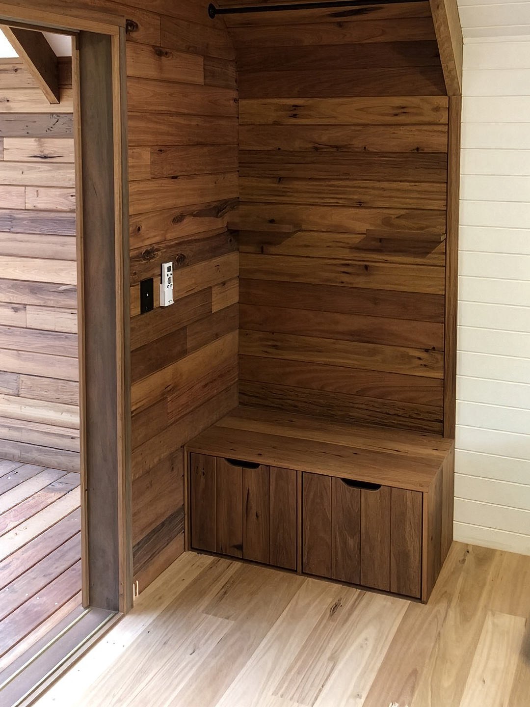 Corner of a room with wood-paneled walls and a built-in bench with storage cabinets. There are electrical outlets and switches on the wall, and a sliding glass door with a wooden frame to the left.