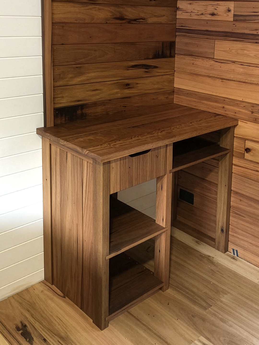 A partially assembled wooden desk with open shelves, set against a wood-paneled wall, with a white shiplap wall on the side and electrical outlets on the wall.