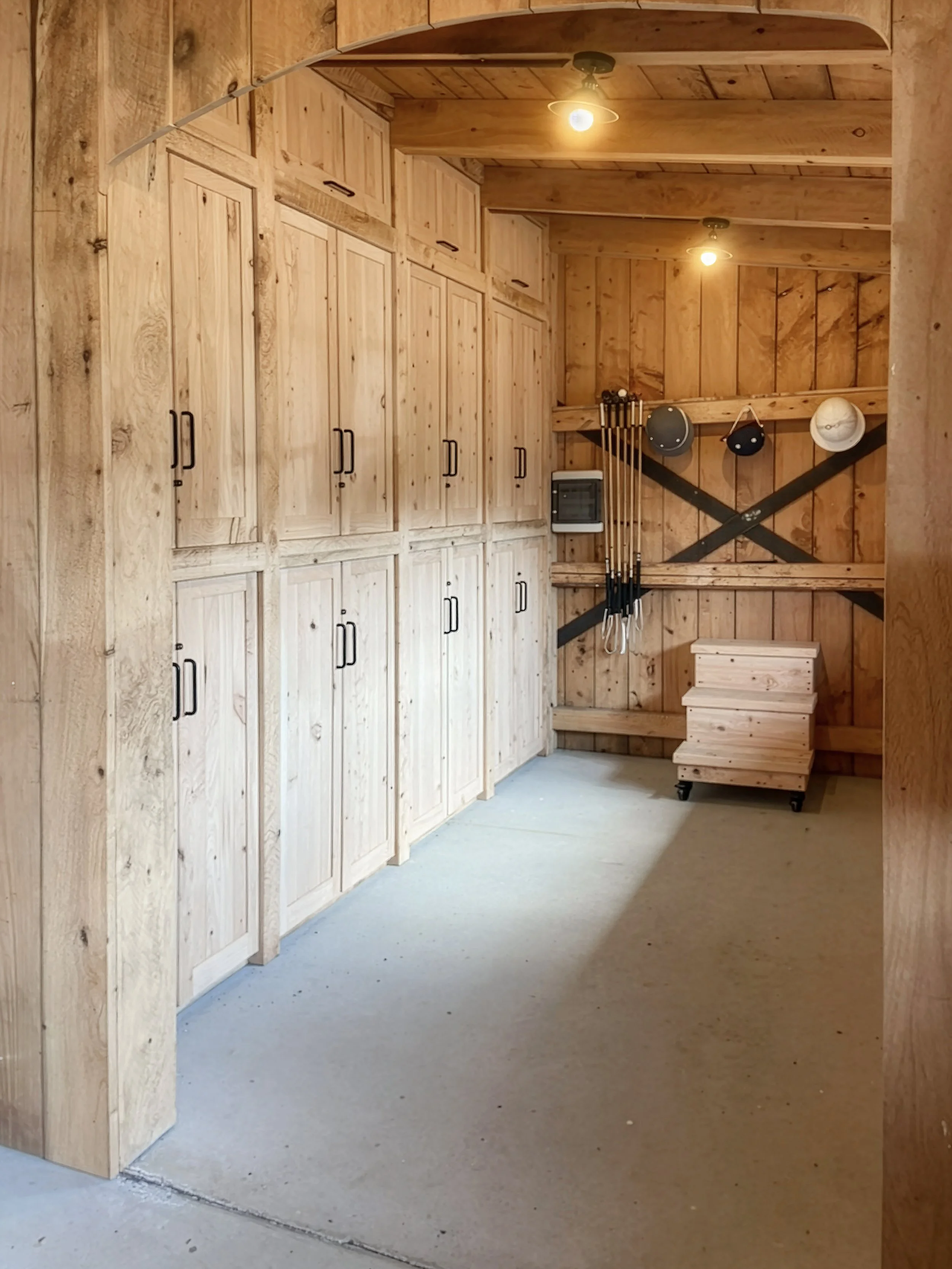 Wooden storage cabinets and a wall with some hooks holding golf clubs and hats. A small heater is mounted on the wall, and a wooden stool with wheels is in the corner.