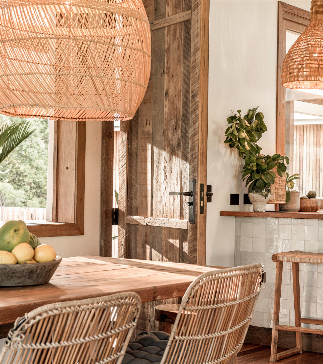 A cozy dining area with a wooden table, rattan chairs, woven pendant lights, and a wooden door. There is a bowl of fruit on the table and a shelf with a green plant and decorative items. Sunlight streams through the window, highlighting the natural wood and greenery outside.