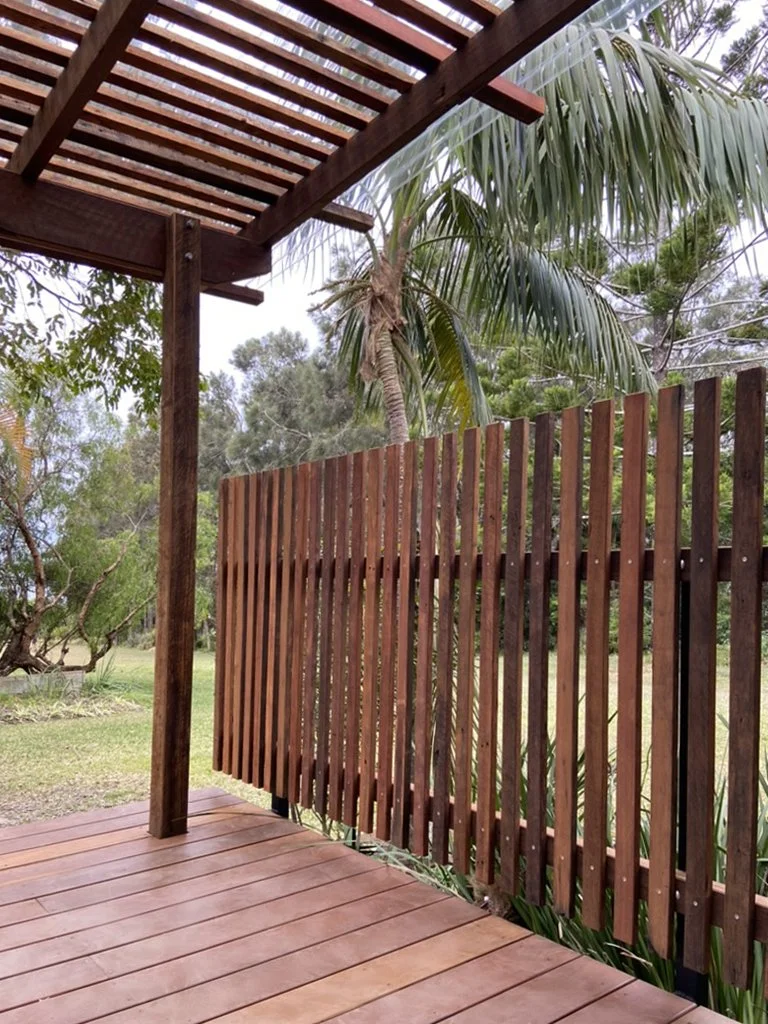 View of a wooden deck with a slatted railing, a wooden pergola overhead, and a background of trees including a prominent palm tree.