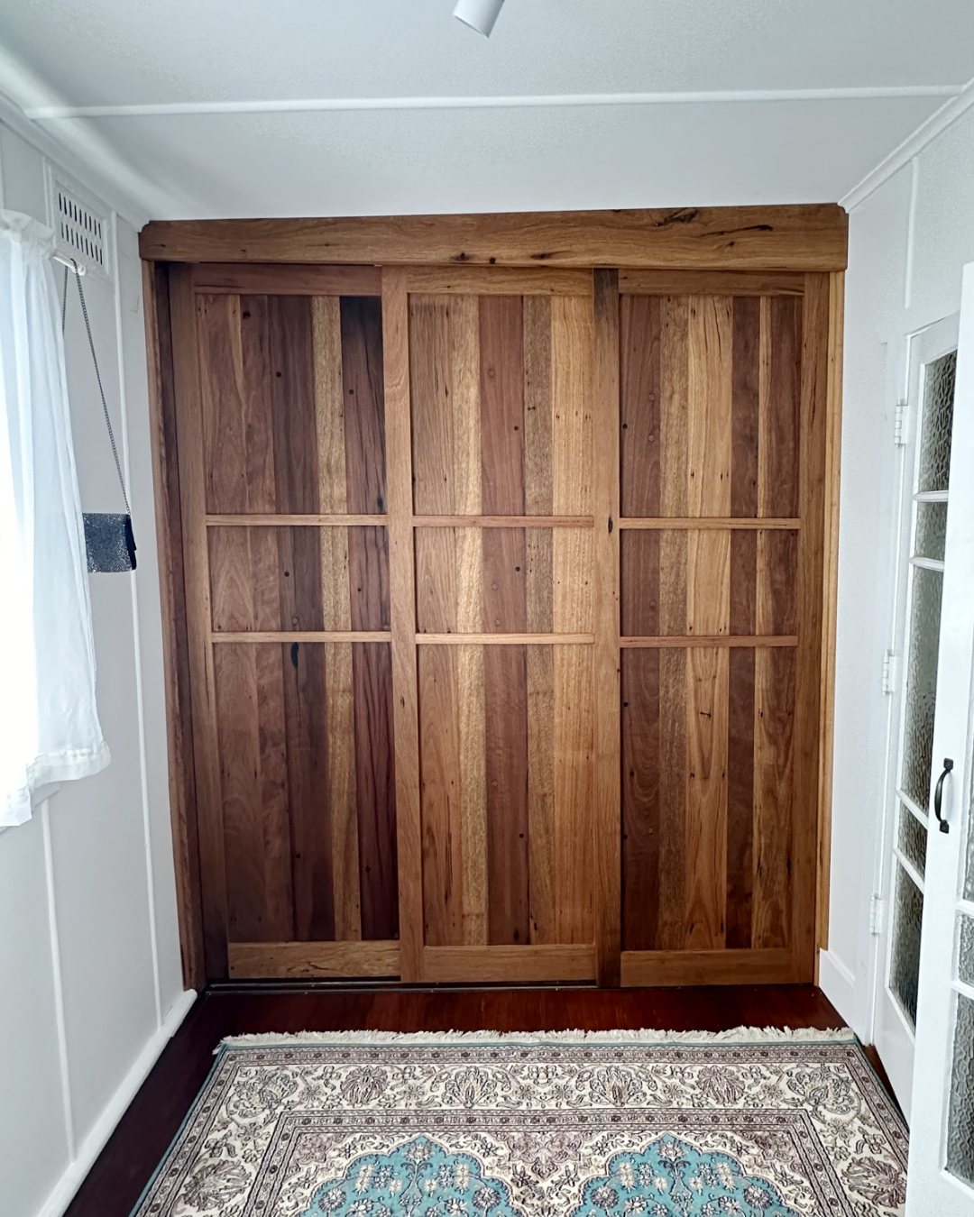 Wooden sliding closet doors with vertical panels in a room with white walls, a patterned rug on the floor, and a window with white curtains.