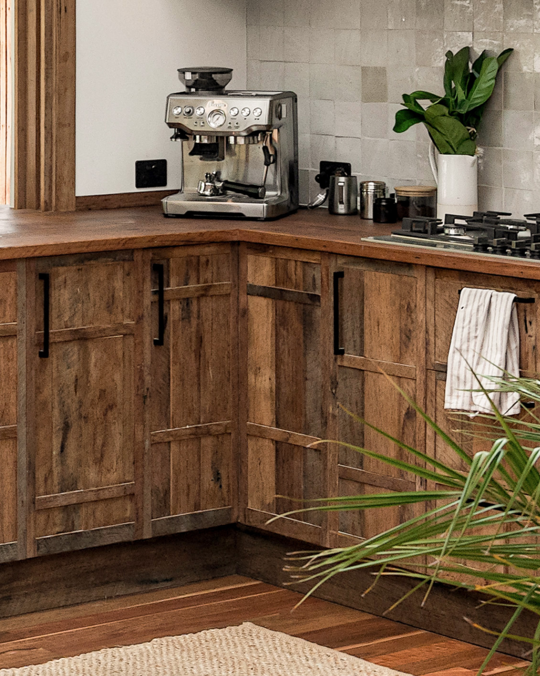 A kitchen with wooden cabinets, a coffee machine on the countertop, a pot with green plants, and a towel hanging on the cabinet handle.