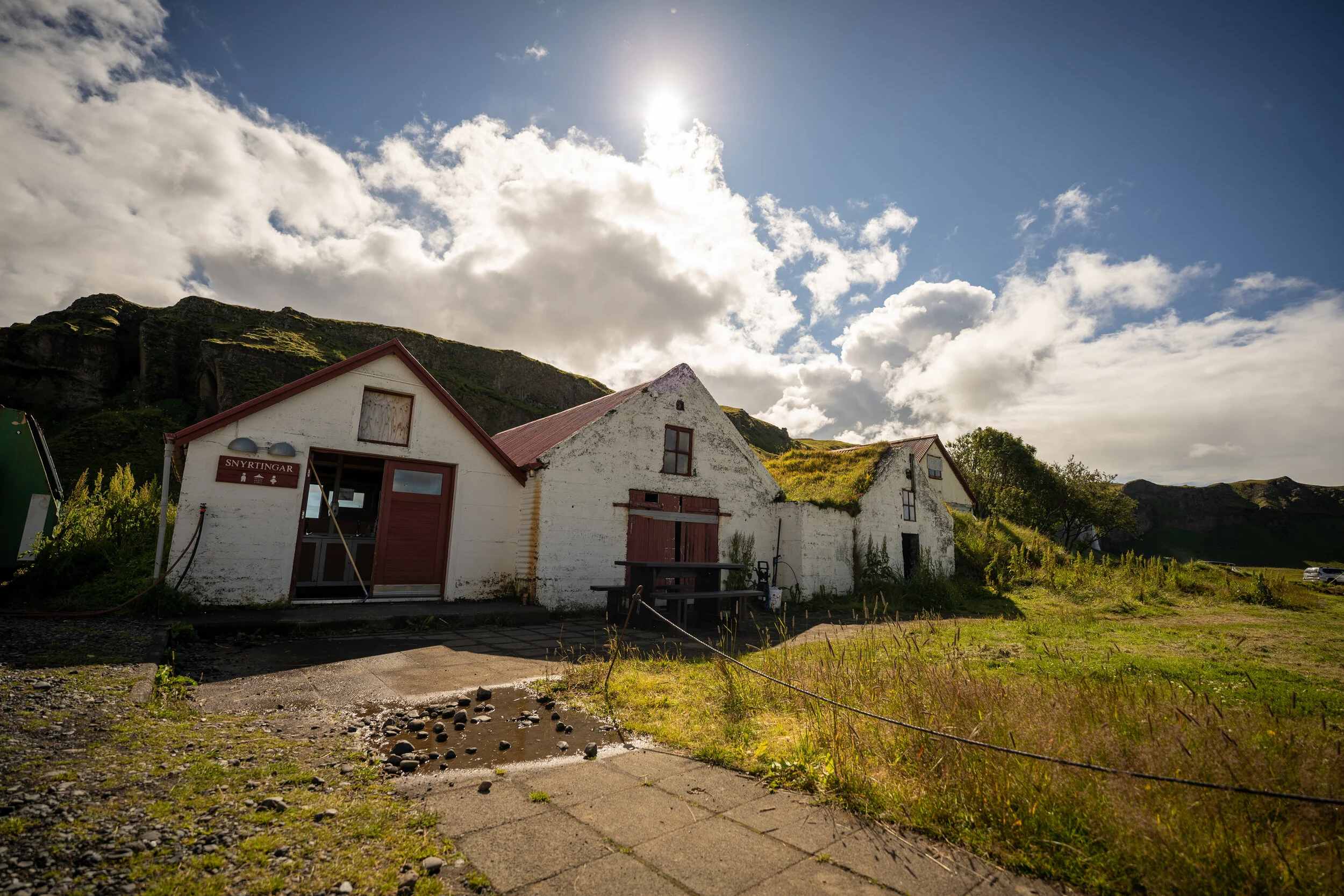 White buildings with red roofs under a partly cloudy sky, with a mountain in the background and some greenery around.