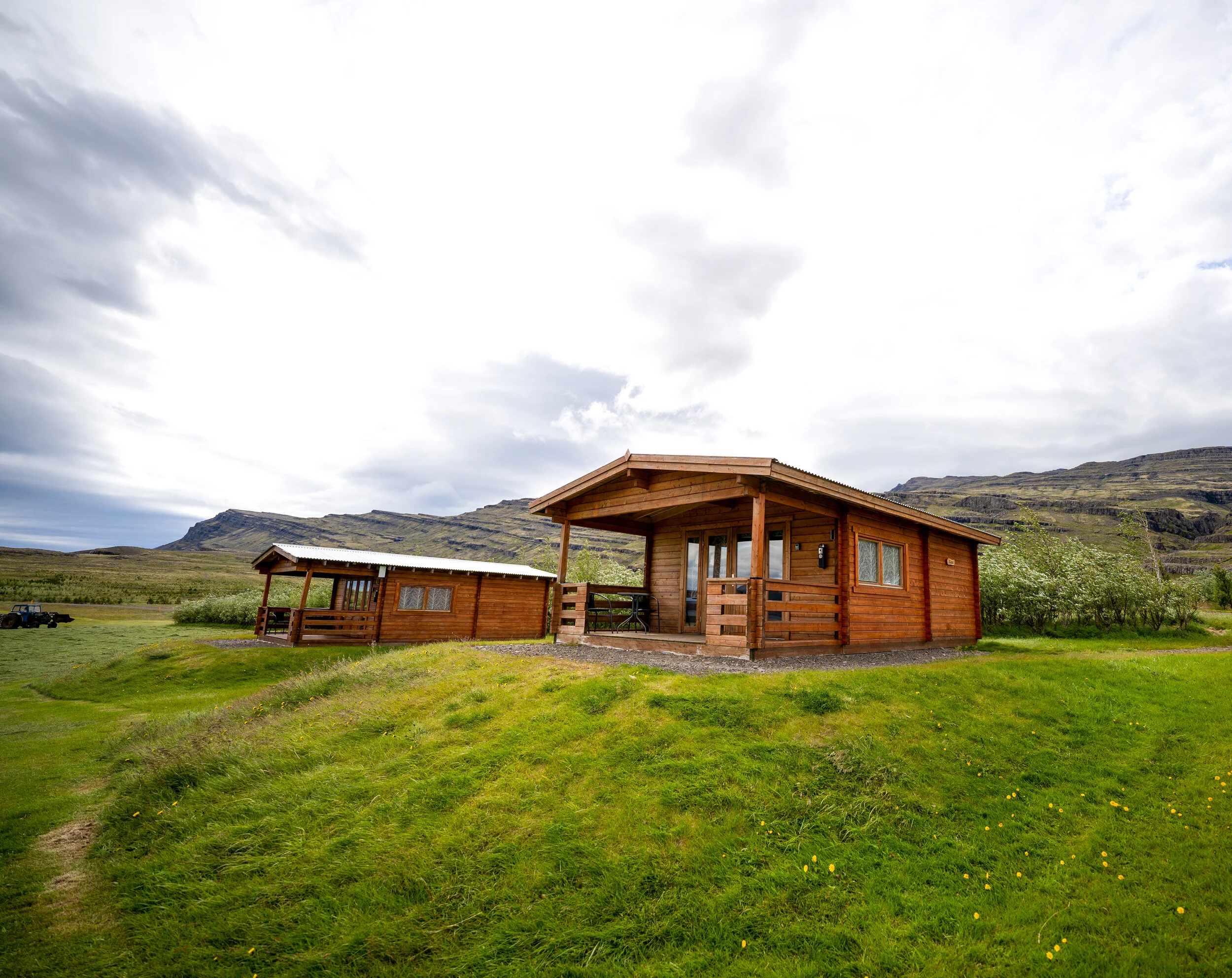 Two wooden cabins on a grassy field with mountains in the background under a cloudy sky.
