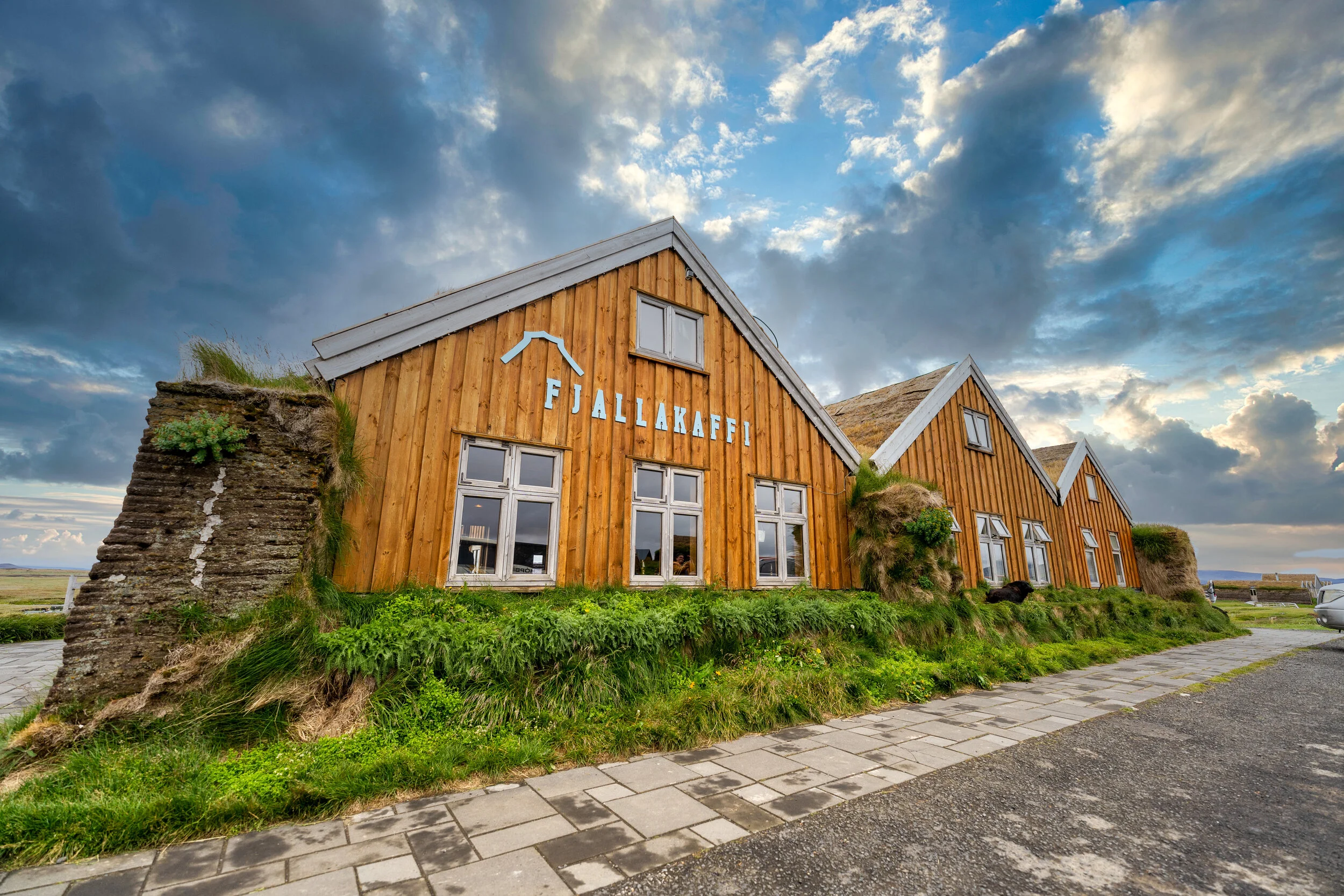 Exterior view of a wooden building with green grass and shrubs in front, under a cloudy sky, with a sign that reads 'Fjallakaffi'.