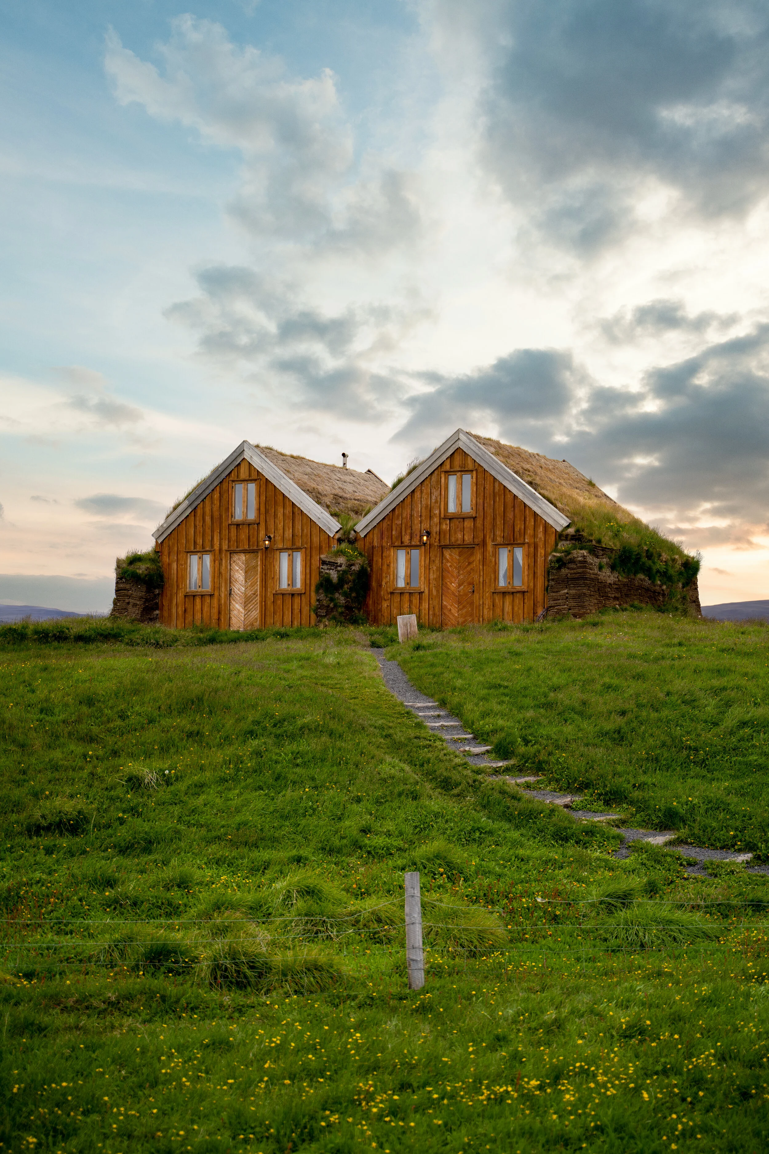 Two traditional turf houses with wooden exteriors and grass roofs situated on a grassy hill under a cloudy sky.