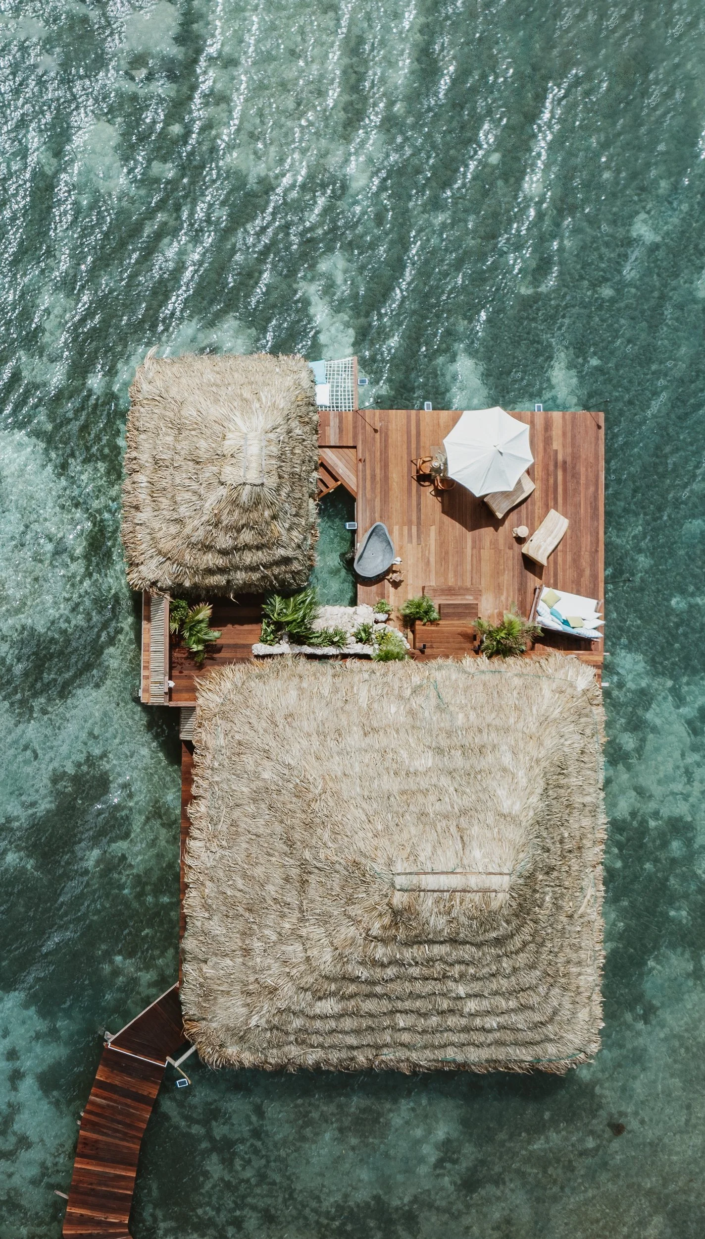 Overhead view of a wooden deck on water with thatched roof structures, outdoor furniture, an umbrella, and potted plants.