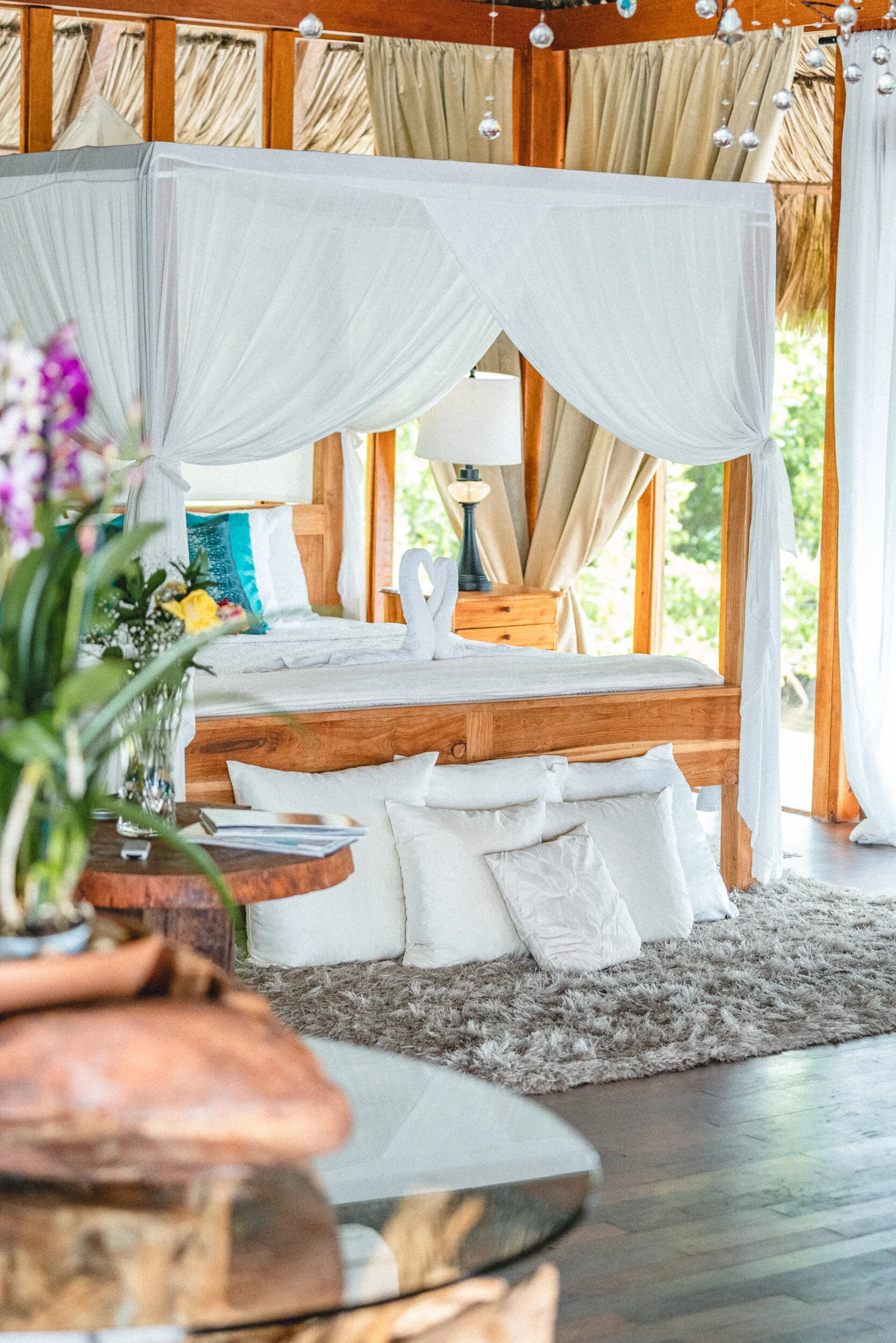 A cozy bedroom with a wooden four-poster bed, white curtains, and swan towel decorations. There are white pillows on the bed and pillows on the floor. A bedside table with a colorful lamp and a window with trees outside in the background.