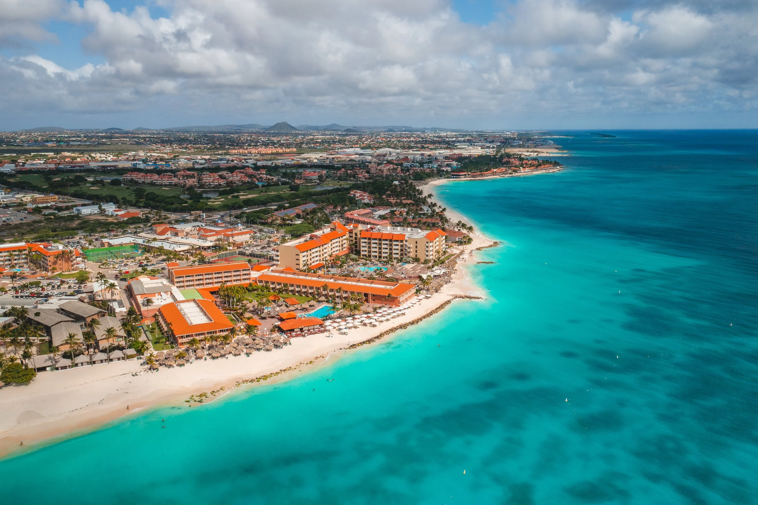 Aerial view of a beachside resort with orange-roofed buildings, white sandy beach, and turquoise ocean waters, with a cityscape and mountains in the distance under a partly cloudy sky.
