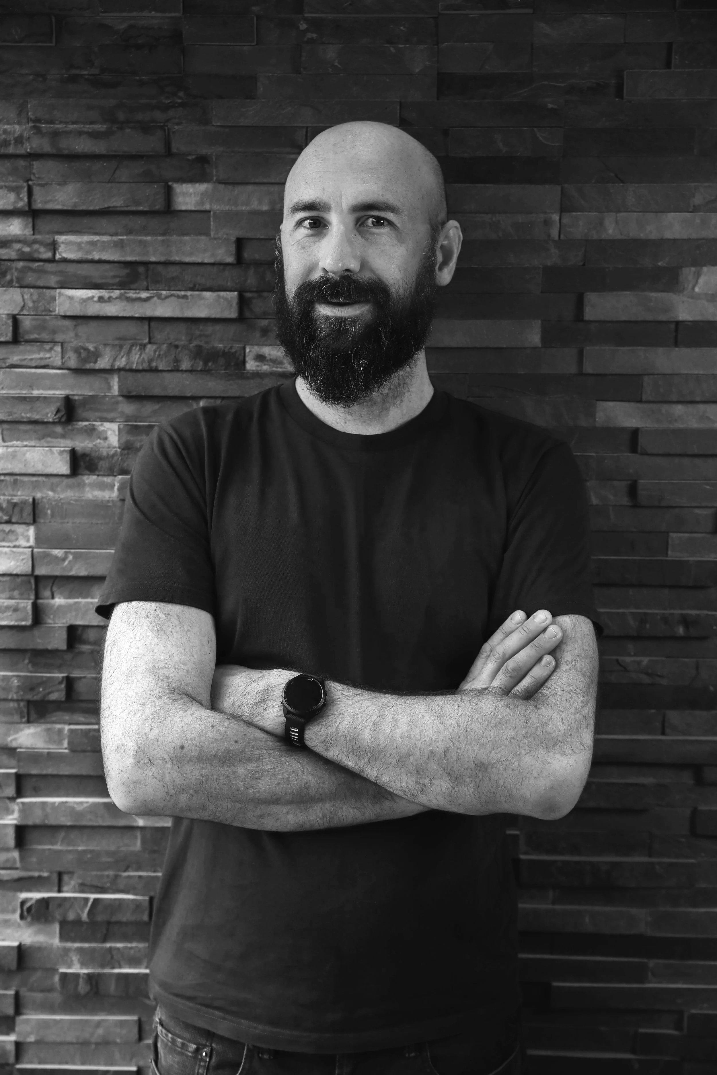 A man with a bald head and a full beard, wearing a black t-shirt and a smartwatch, crossing his arms and smiling slightly while standing against a textured brick wall.