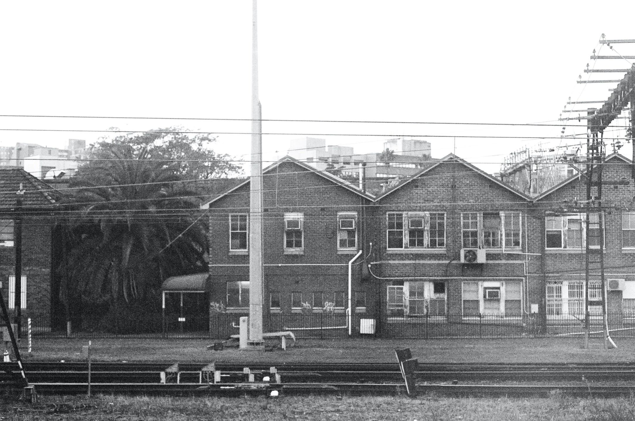 Black and white photo of a residential building with multiple windows, surrounded by power lines and trees, next to a railway track.
