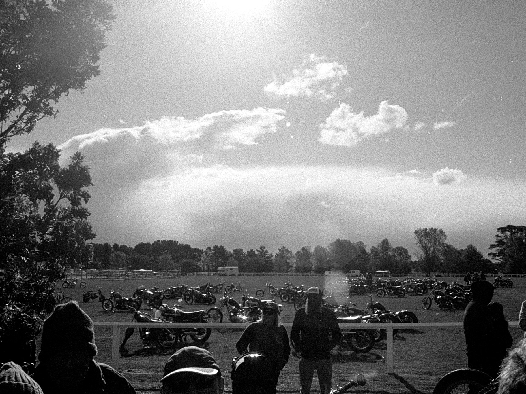 A black and white photo of a park with motorcycles lined up in the background, people standing in the foreground, trees on the sides, and a partly cloudy sky overhead.