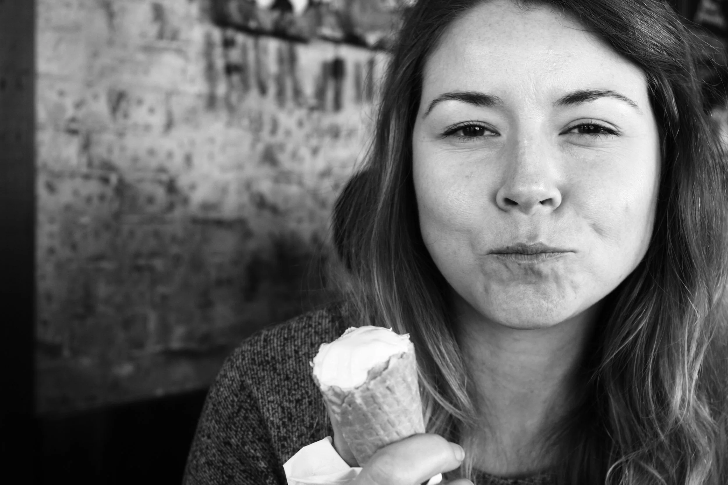 A woman with long hair holding an ice cream cone, making a playful facial expression, in front of a brick wall, in black and white.