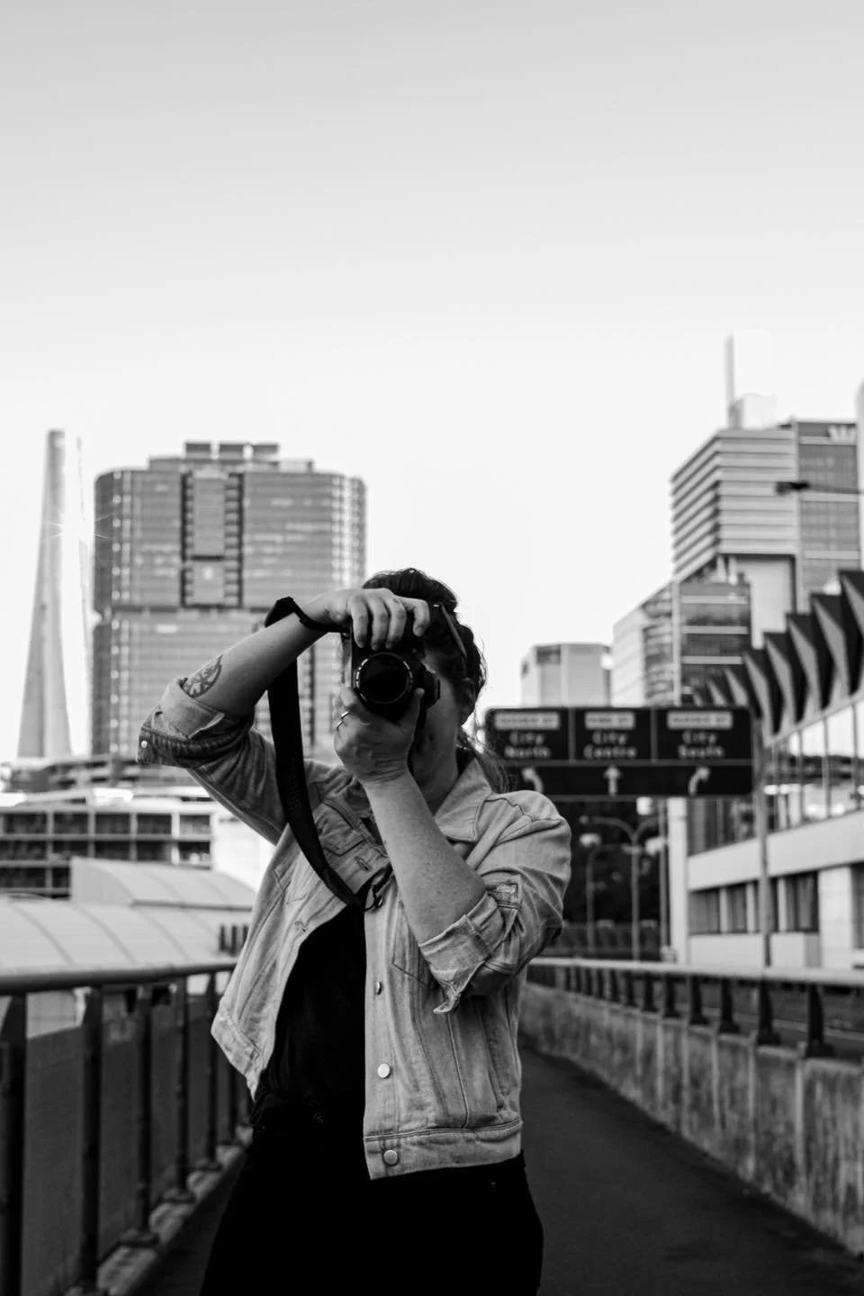 A person taking a photo with a camera on an urban rooftop or bridge with city buildings and skyline in the background.