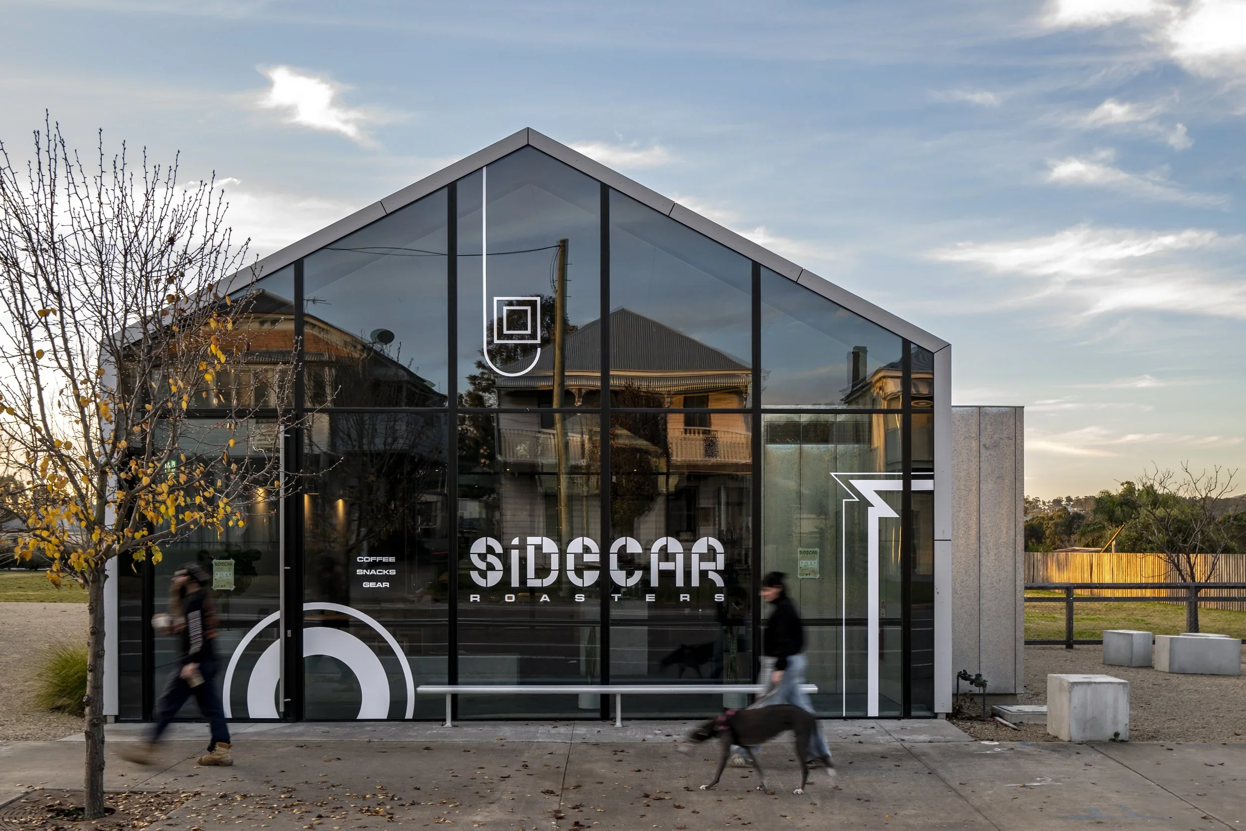 Modern glass-roofed coffee shop called Sidecar Roasters with people walking past, one person with a dog, during sunset in a suburban area.