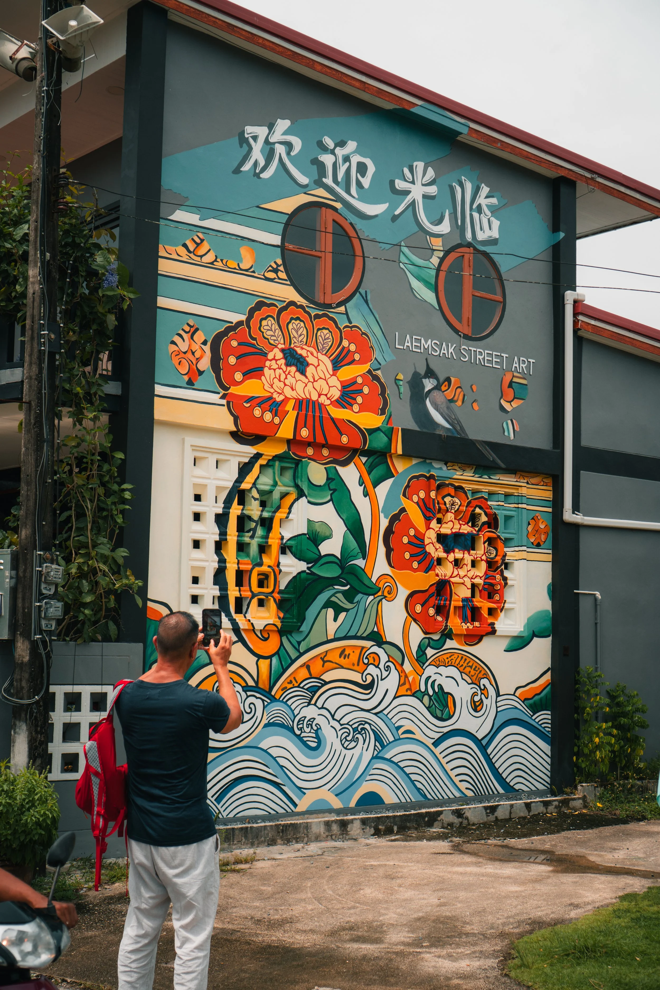 A color mural painted on a building in Laem Sak, Thailand, with a visitor in the foreground taking a picture of the mural with their phone.