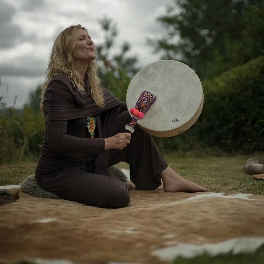 A woman sitting on the ground outdoors on a blanket, playing a large frame drum with a decorated drumstick in her hand, with trees and cloudy sky in the background.