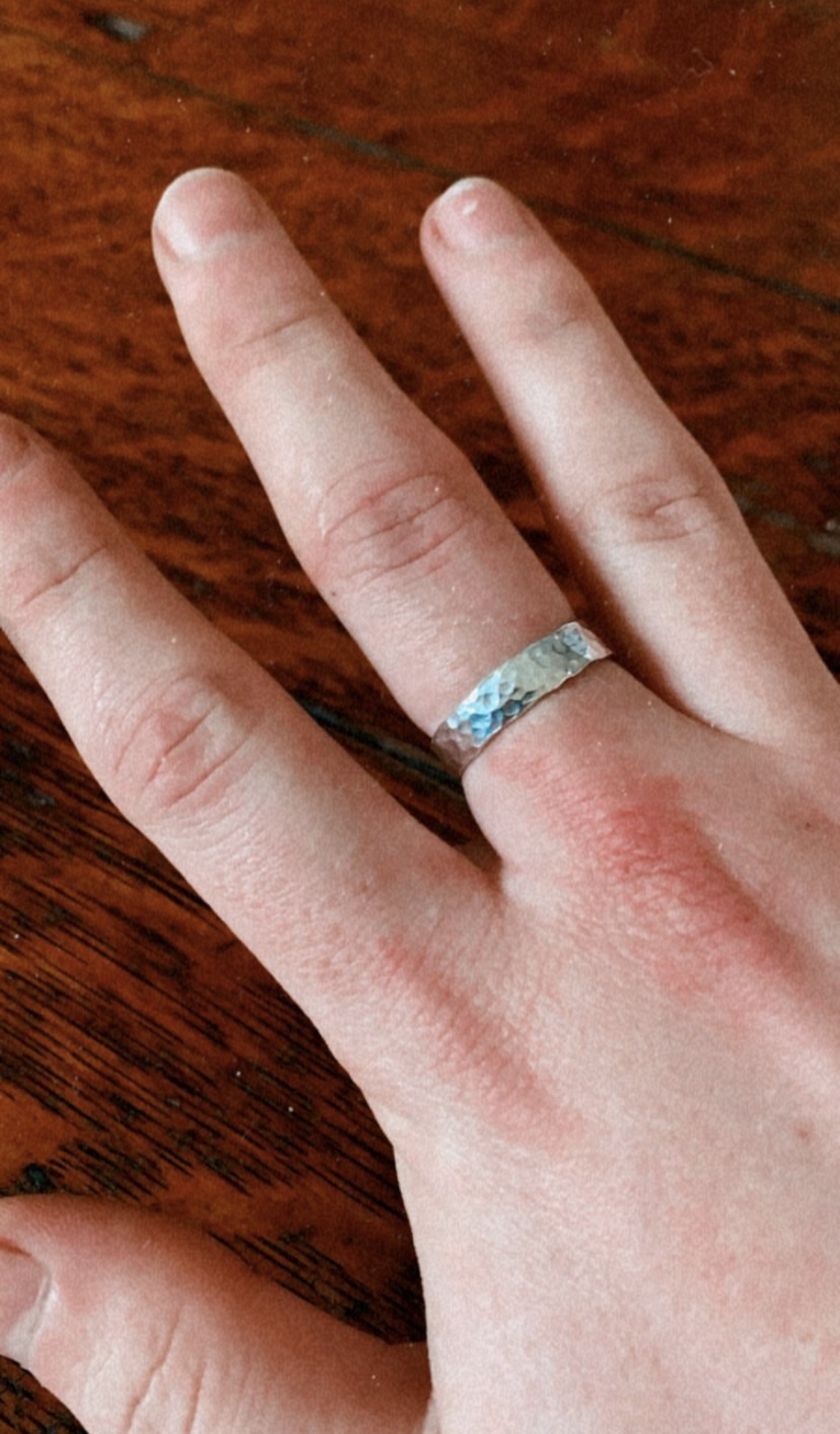 Close-up of a hand with fair skin, showing a silver ring with a hammered texture on the ring finger, resting on a wooden surface.