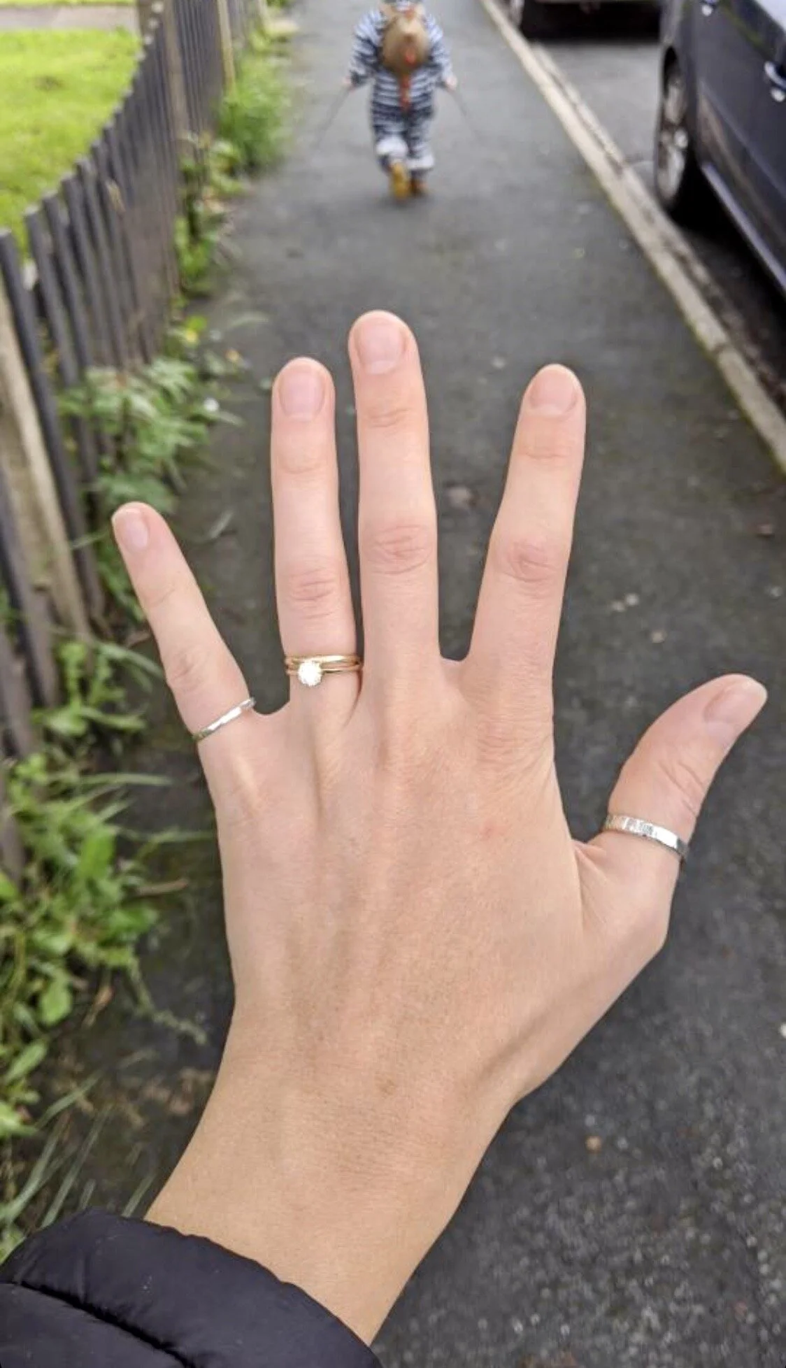 Close-up of a person's left hand with rings on their fingers, reaching out toward a sidewalk. In the background, a young child in striped pajamas is walking away, holding a leash.