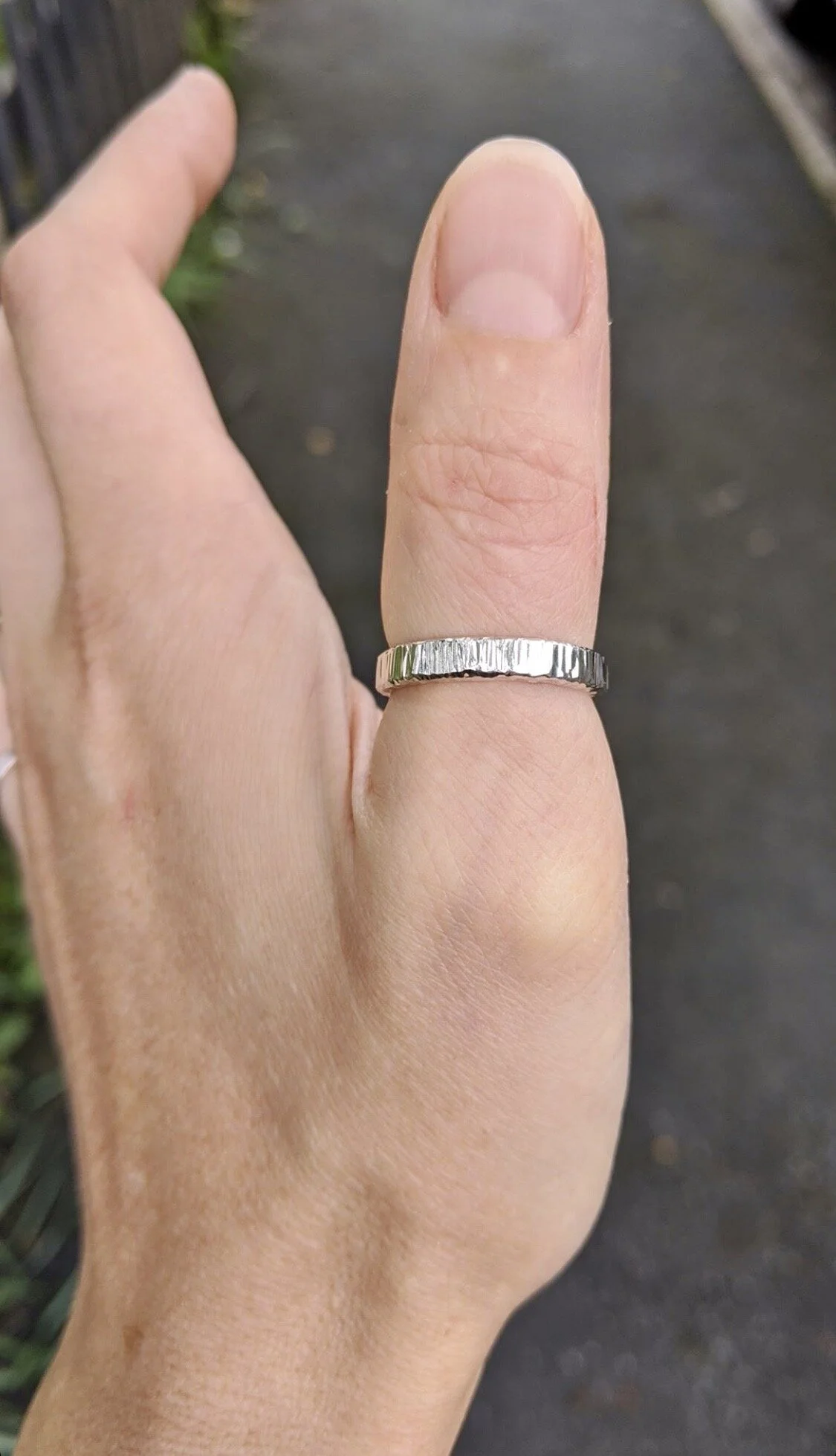 Close-up of a hand wearing a silver textured ring on the middle finger, outdoors on a dark pathway.