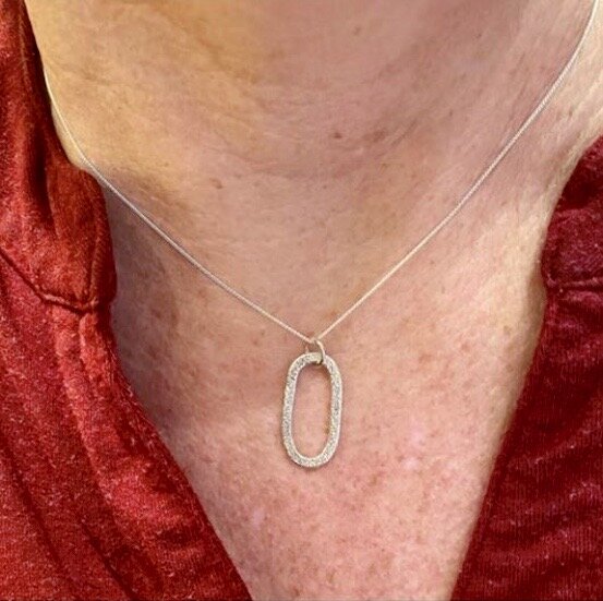 Close-up of a person's neck wearing a silver necklace with an oval-shaped pendant encrusted with small diamonds, and a red clothing garment.
