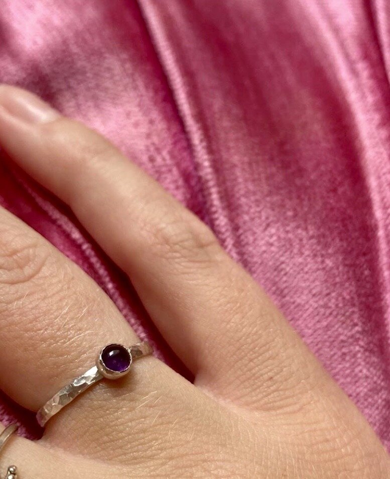 A close-up of a hand wearing a silver ring with a small, round, dark purple stone on a finger, with a pink, textured fabric in the background.