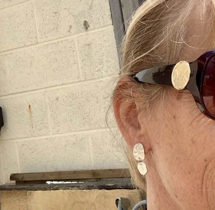 Close-up of a woman wearing black sunglasses with a gold emblem on the side and dangling earrings, against a background of a beige cinder block wall.