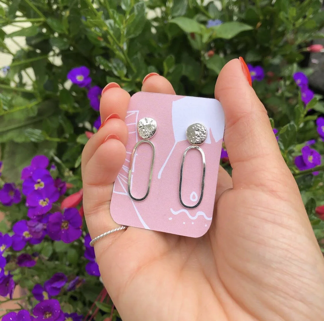 Hand holding a pair of stylish earrings with a dark purple flower background.