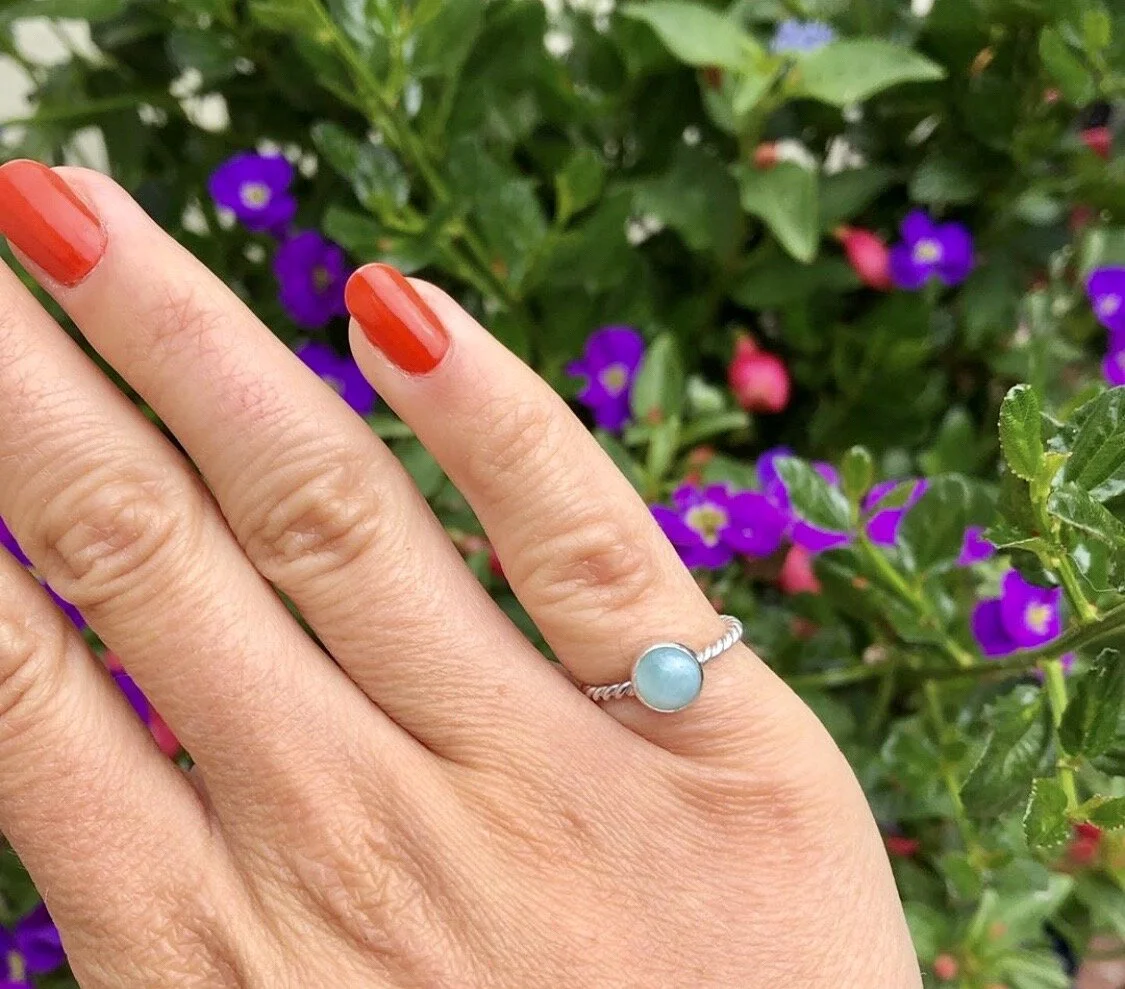 A hand with red polished nails wearing a ring with a light blue gemstone, against a background of purple and pink flowers and greenery.