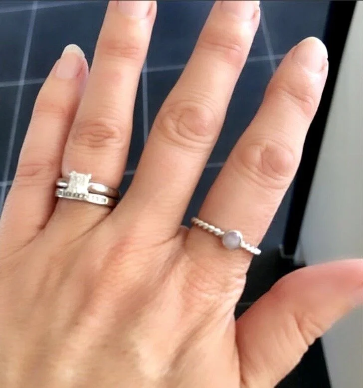 A person's hand showing three rings: a large engagement ring with a central diamond, a plain wedding band, and a delicate band with a pearl.