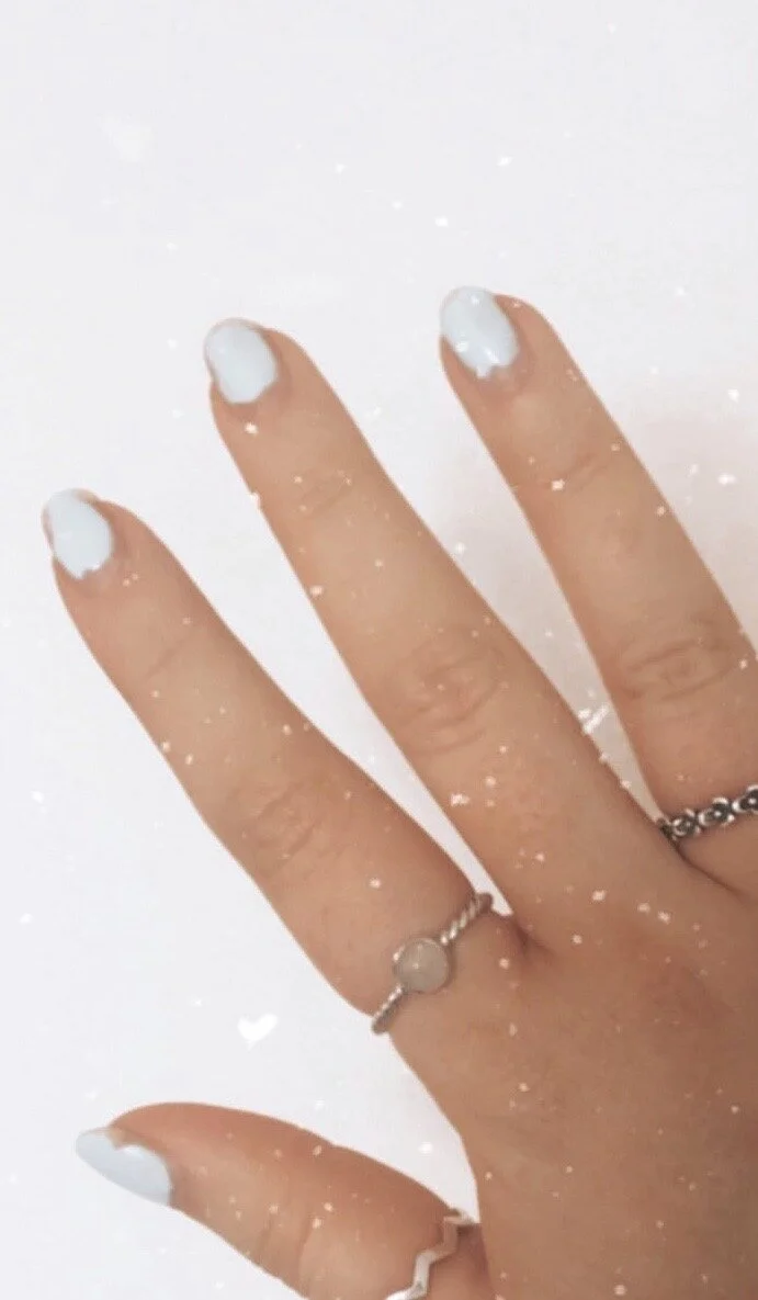 Close-up of a hand with white painted nails, wearing a silver ring with a round stone, against a white background with small white speckles.