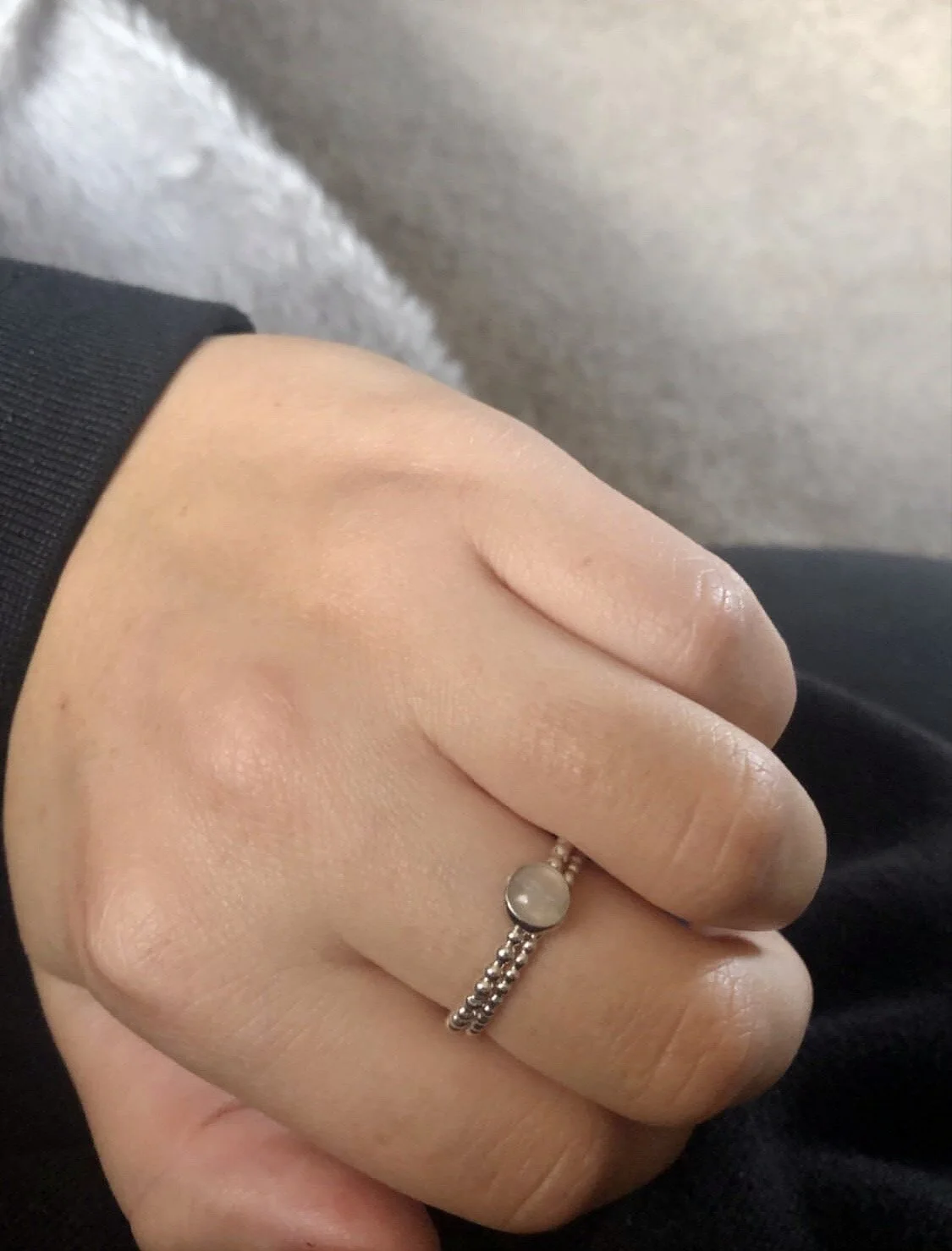 A close-up of a hand wearing a silver ring with a round, milky white gemstone. The hand is resting on a gray textured surface, and the person is wearing a black long-sleeve shirt.