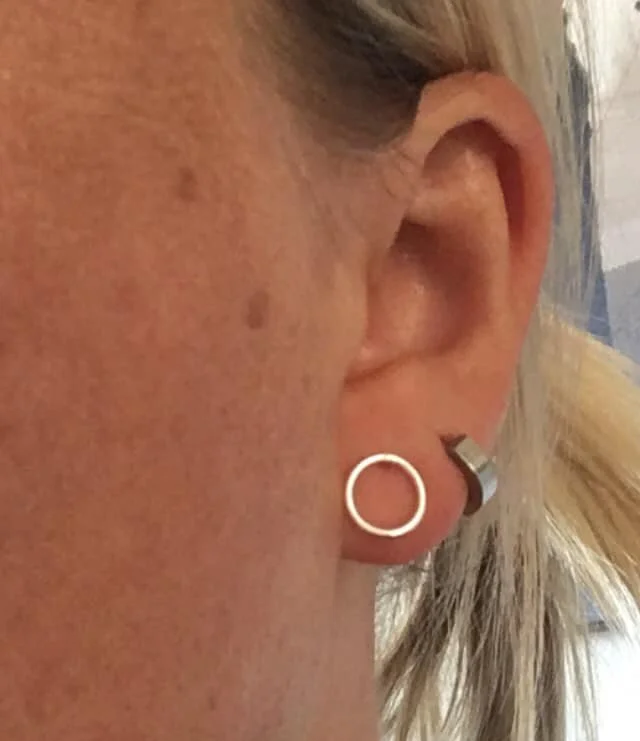 Close-up of a person's ear with a silver hoop earring and a silver conch ring in the cartilage, showing skin with freckles and light-colored hair.