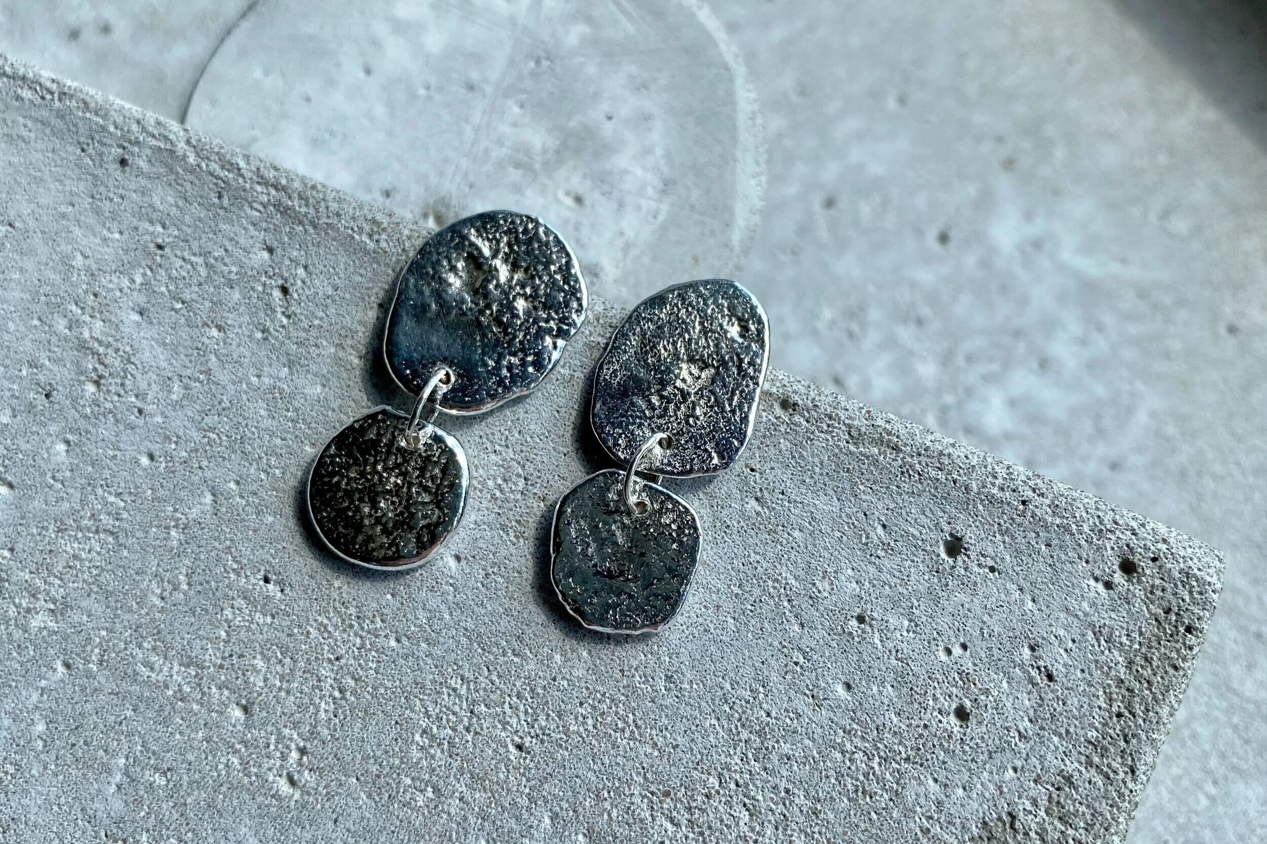 Pair of silver earrings with textured oval and irregular shapes, resting on a concrete surface.