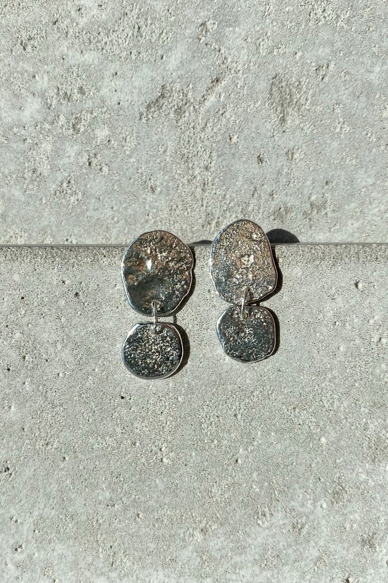 Silver earrings with textured, irregularly shaped oval and circular pendants on a textured, light-colored surface.