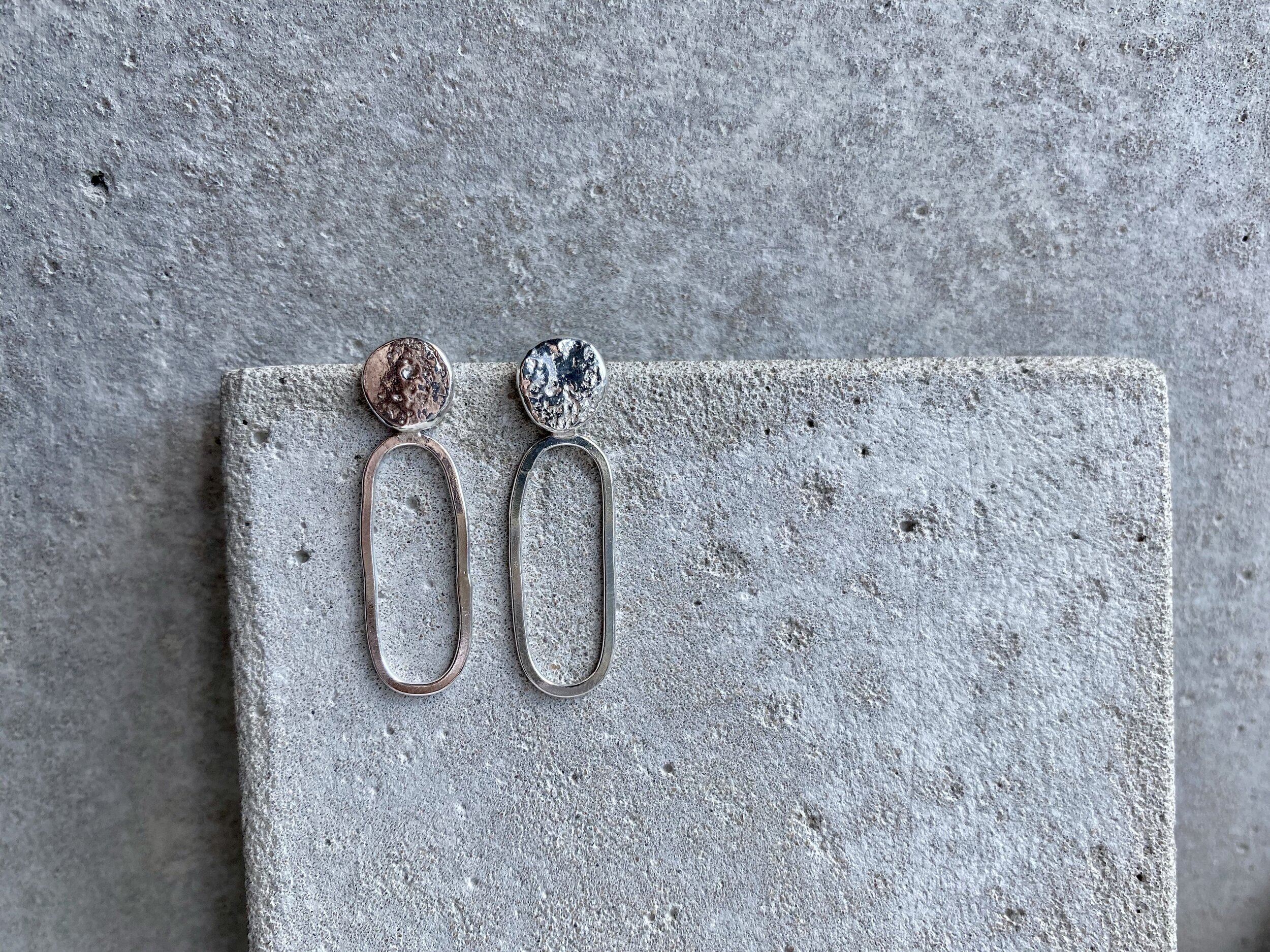 Pair of modern earrings with oval loop design in silver, displayed on a textured gray concrete surface.