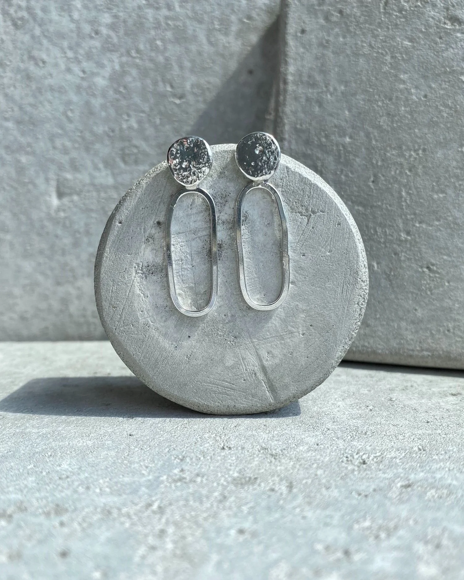 Silver earrings with textured round tops and elongated oval dangles, displayed on a gray round object against a gray background.