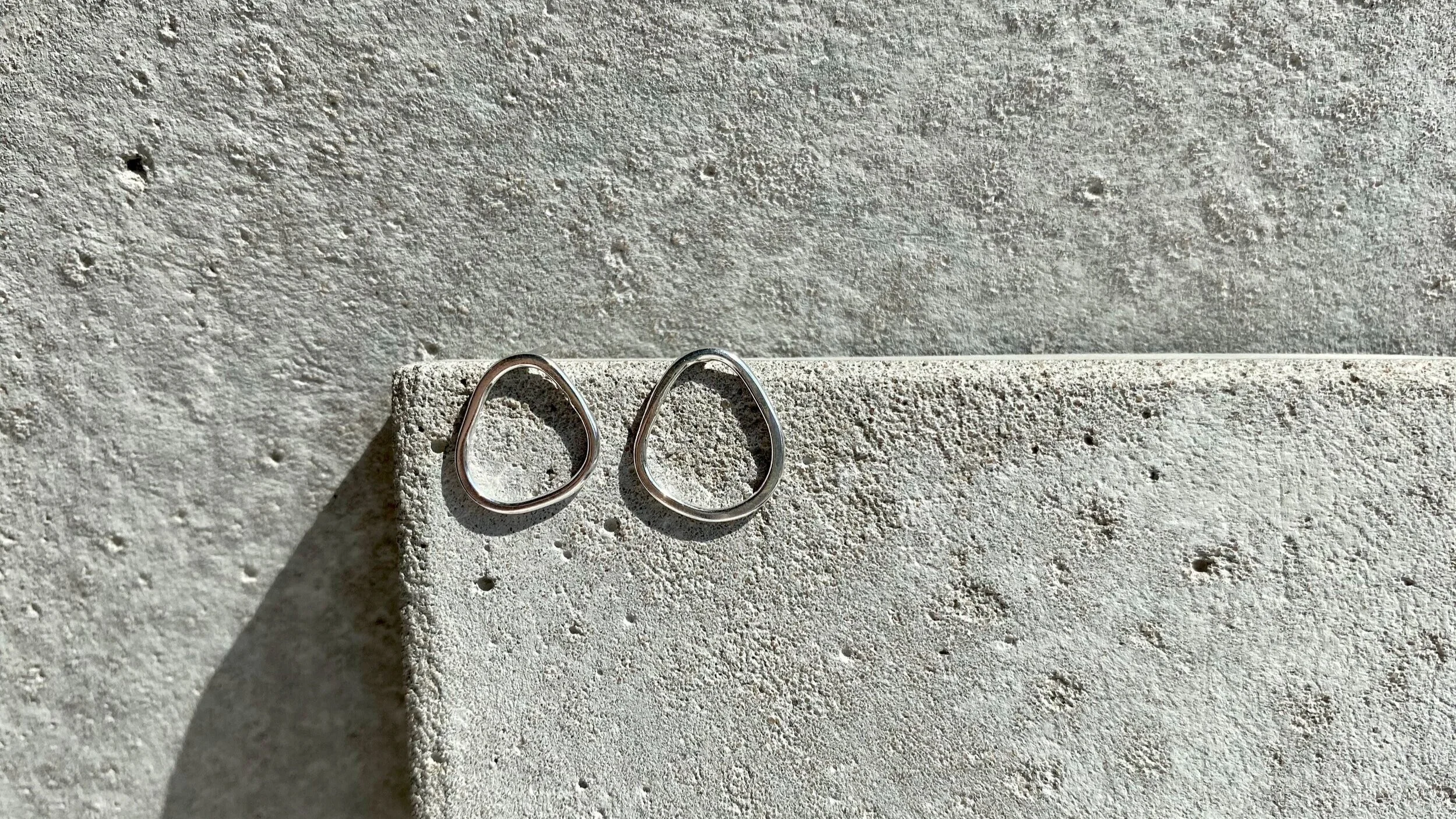 Two silver ring-shaped earrings resting on a light grey textured surface with a shadow cast to the left.