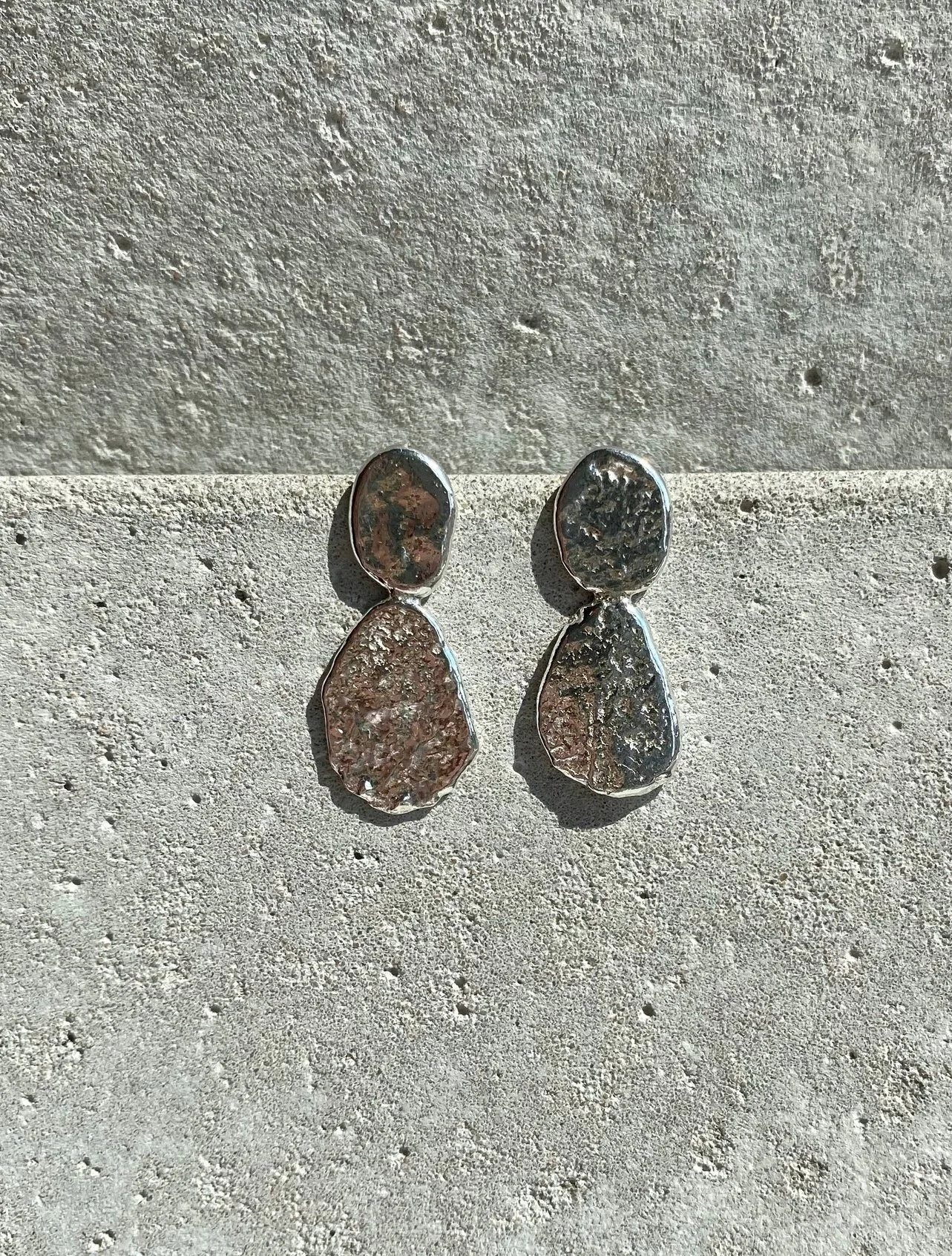 Pair of earrings with irregularly shaped, textured metallic stones on a cement surface.