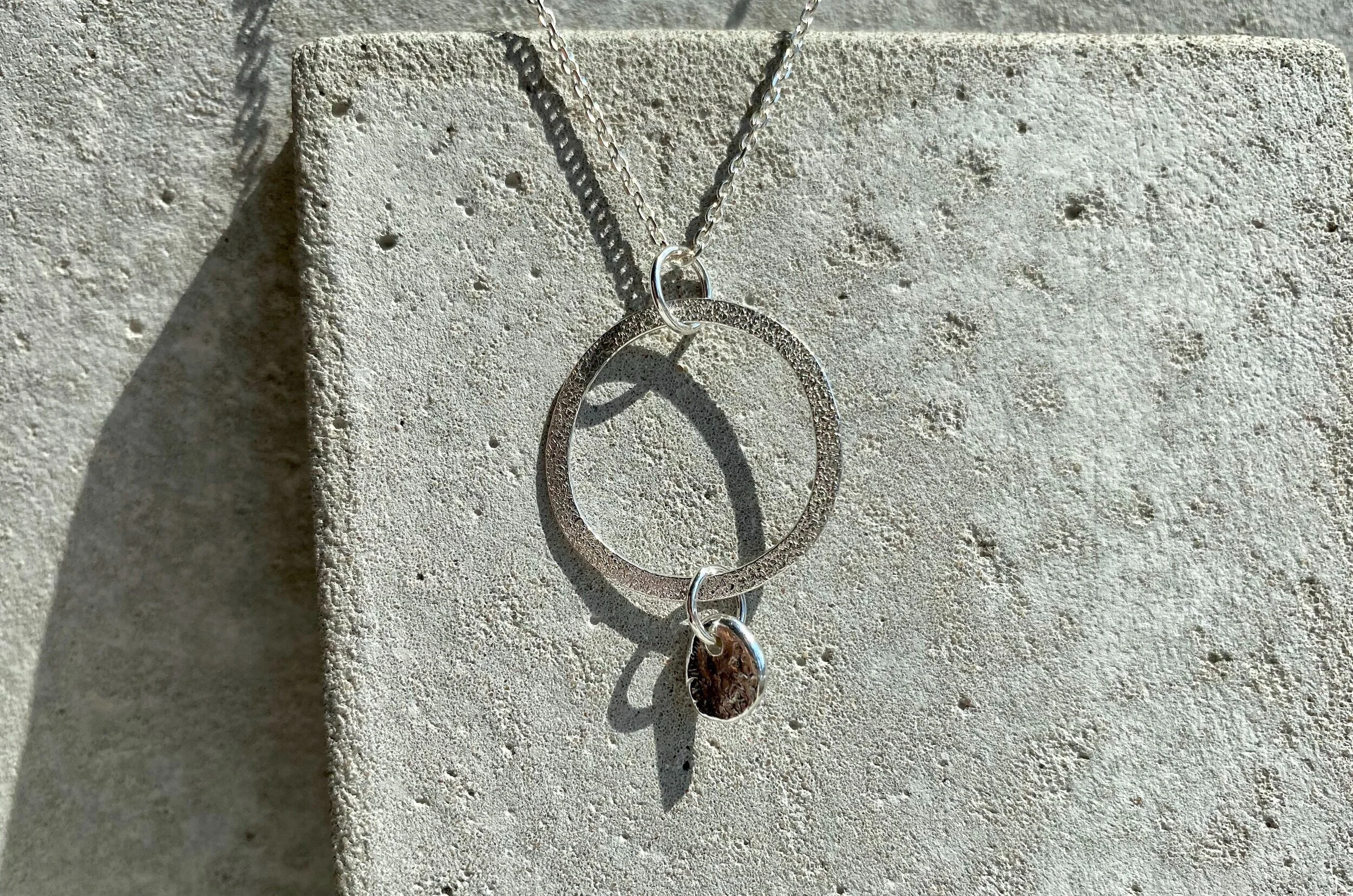 A silver necklace with a circular pendant and a small teardrop-shaped charm hanging from the bottom, placed on a textured, light-colored stone surface.